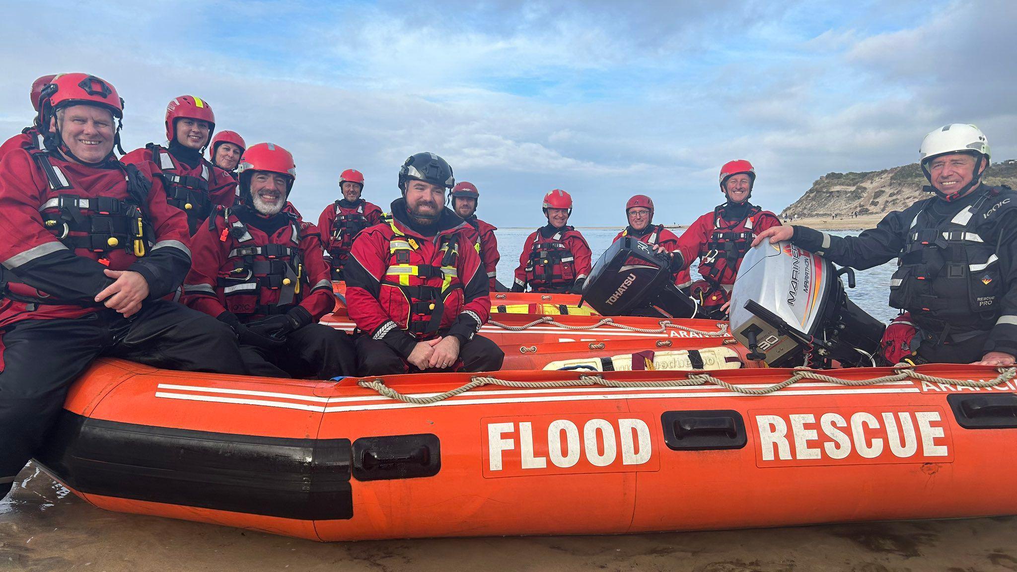 A group photo of 12 of the Cornwall Surf Live Saving club arranged around an inflatable boat on a beach that has 'FLOOD RESCUE' printed on its side. The club members are wearing full kit including buoyancy aids and helmets.