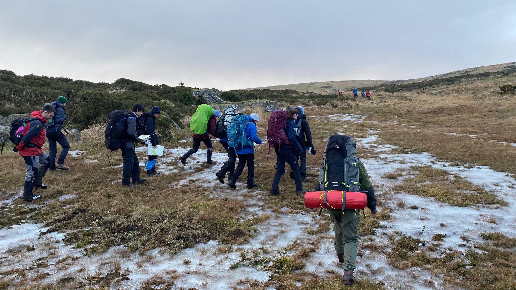 A group of people with rucksacks trekking over the moor which has patches of snow on it.