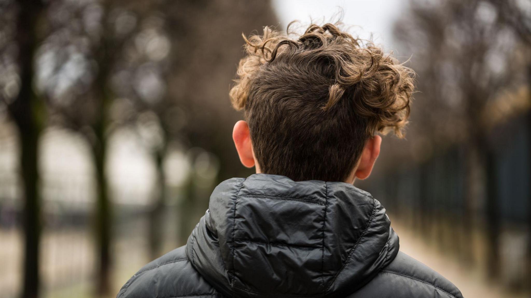 a teenager, with short curly dark hear and wearing a black jacket is facing away from the camera