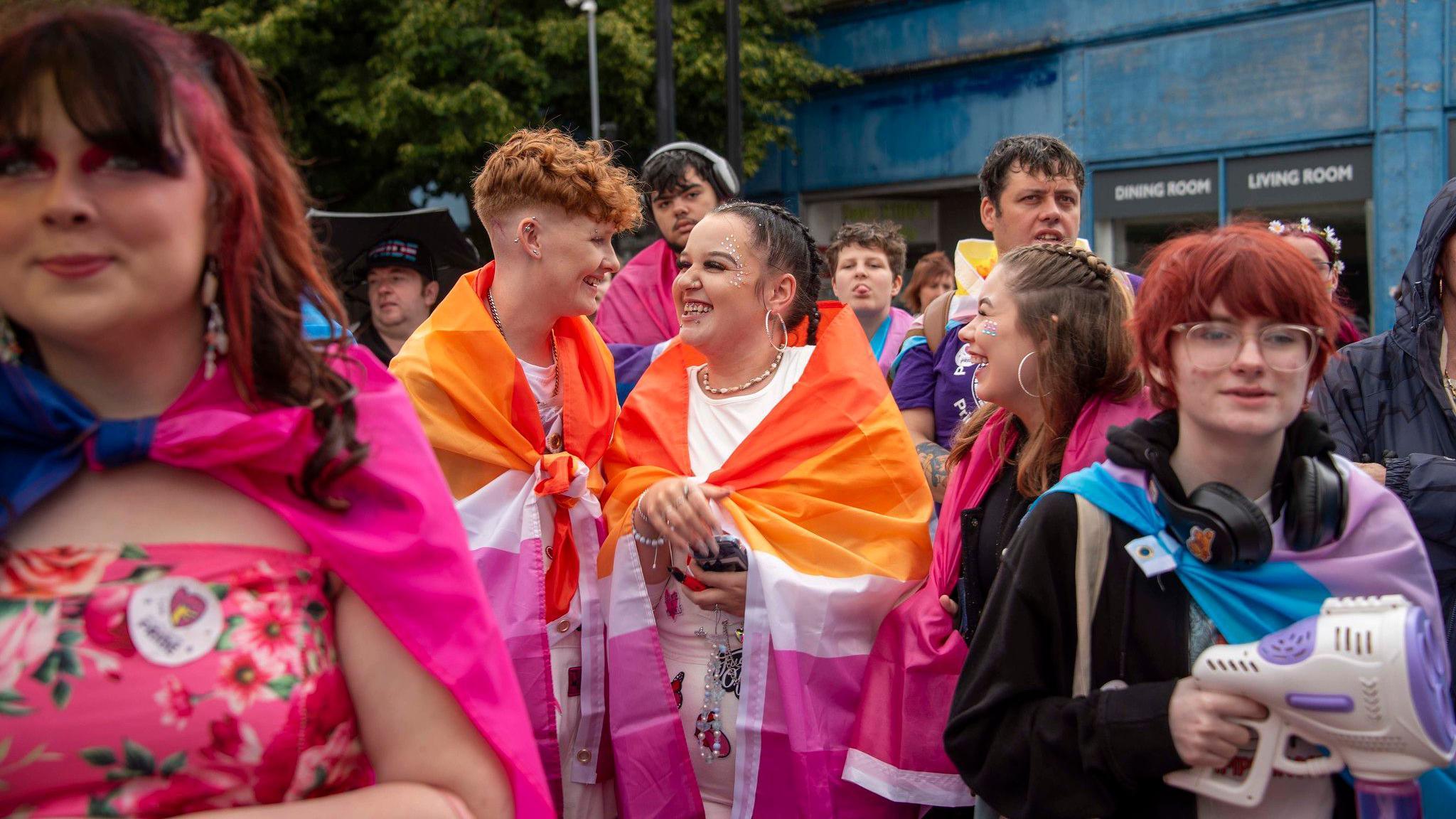 Three people wrapped in large, orange and pink flags, smile at each other while in the middle of a parade. Other people are standing around them.