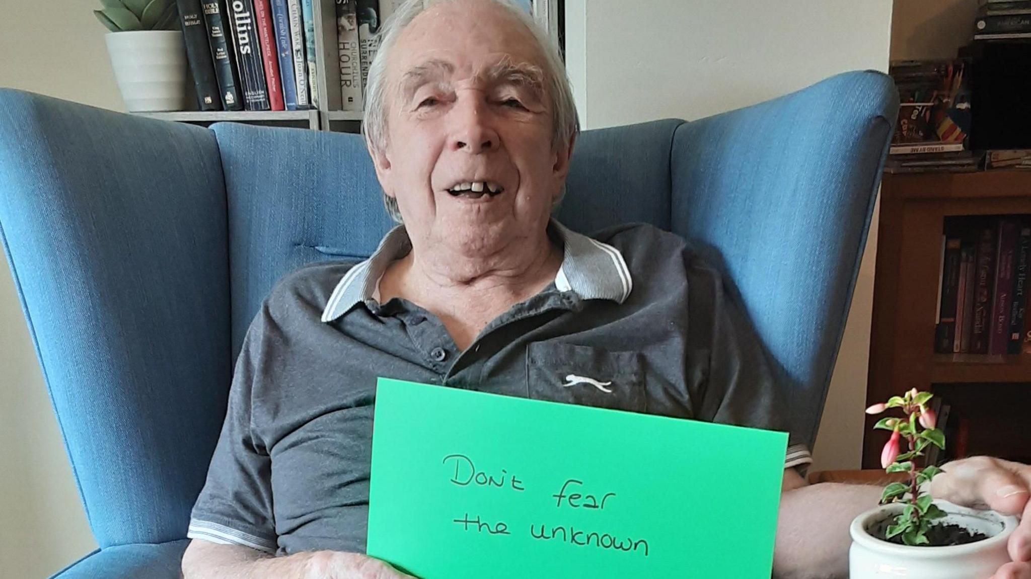 Ken Lewis, 81, sits in a blue armchair smiling at the camera. He has a small plant in one hand and in the other, he is holding a green piece of paper which has handwritten on it 'don't fear the unknown'.