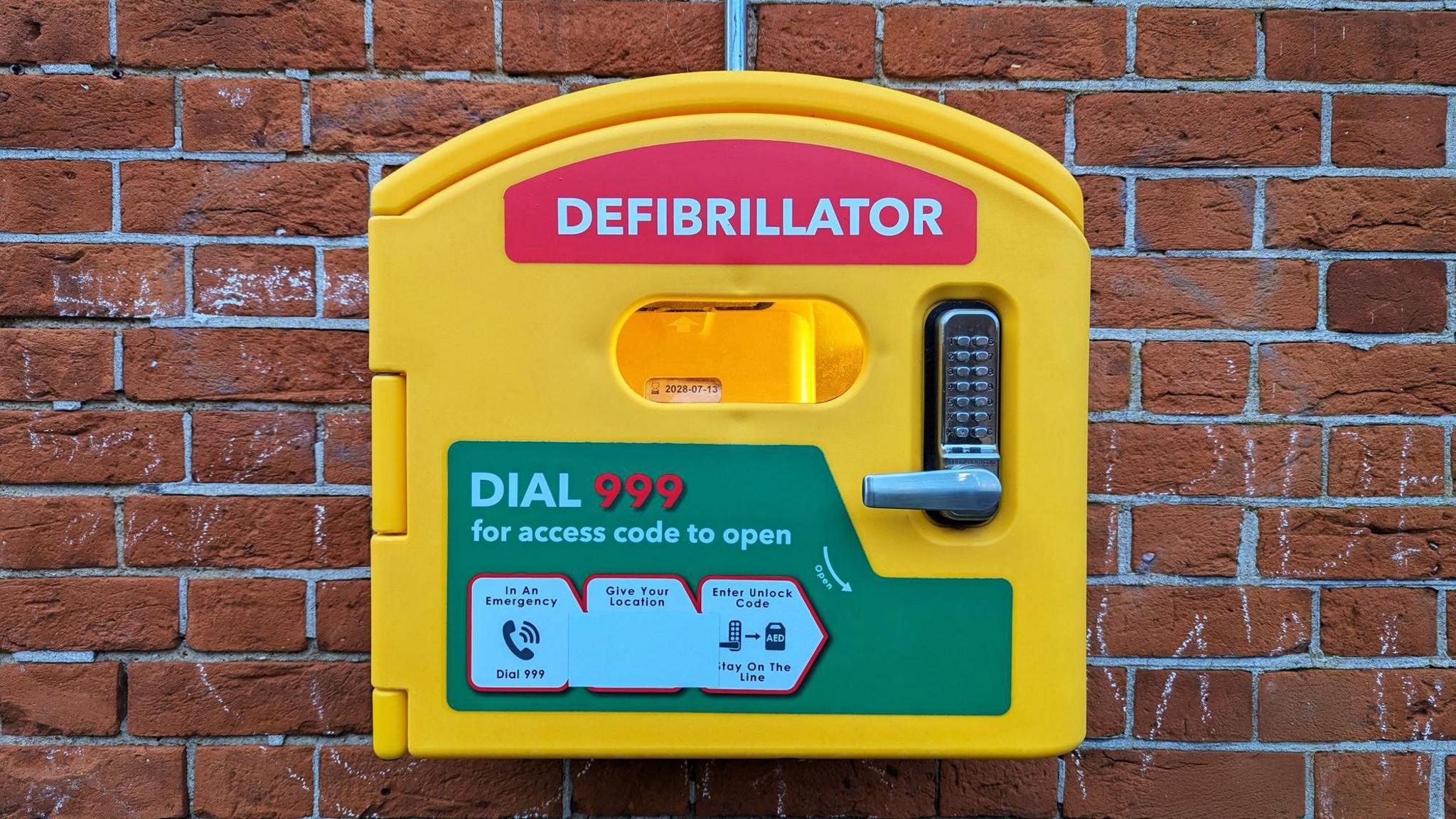 A yellow box defibrillator mounted on a brick wall. It has a red sticker on it that reads, "Defibrillator". There is a keypad and handle, and a green sticker that reads, "Dial 999 for access code to open".