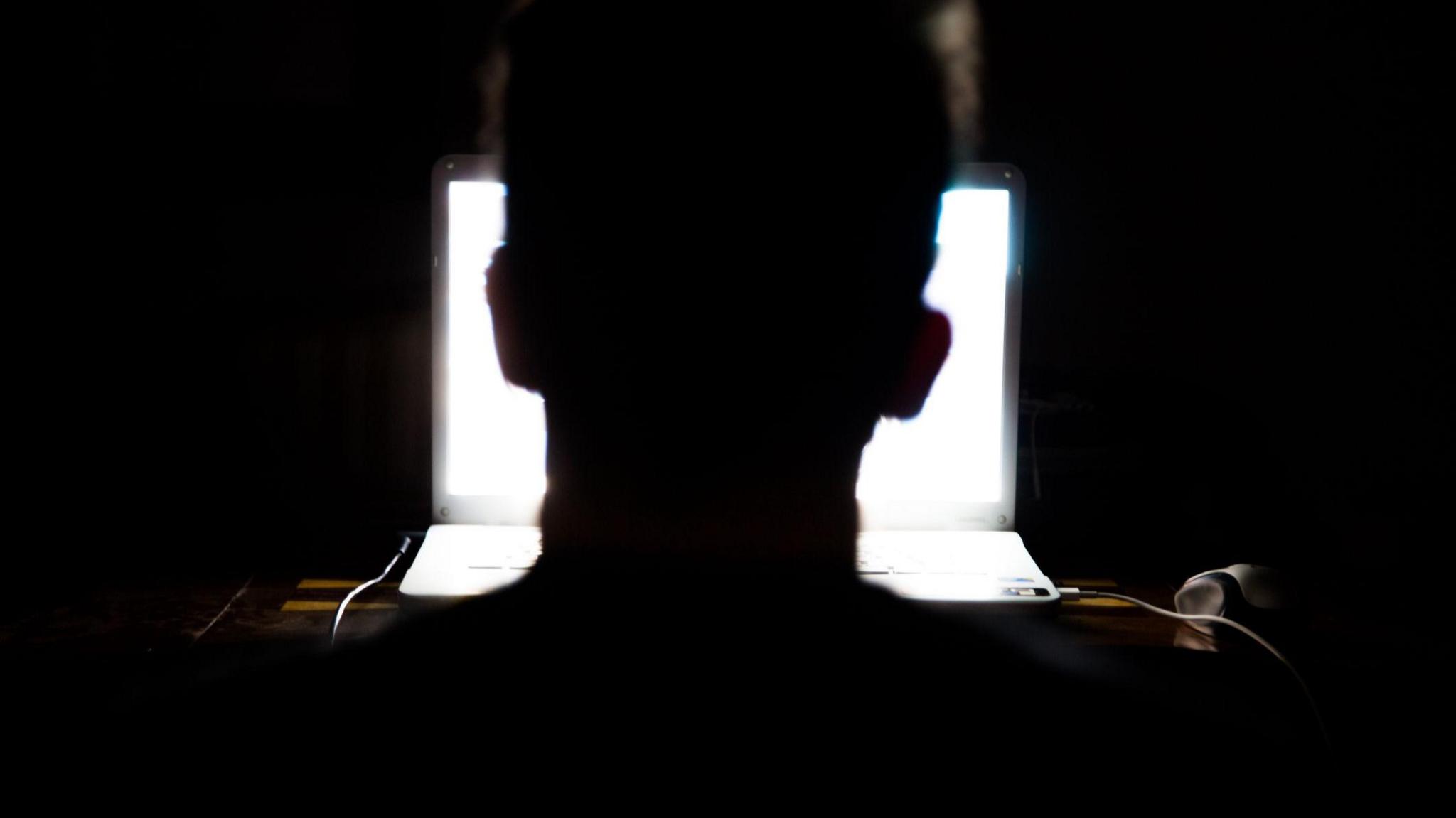A man sits in front of a computer in the dark, with his silhouette illuminated by the light of the screen.