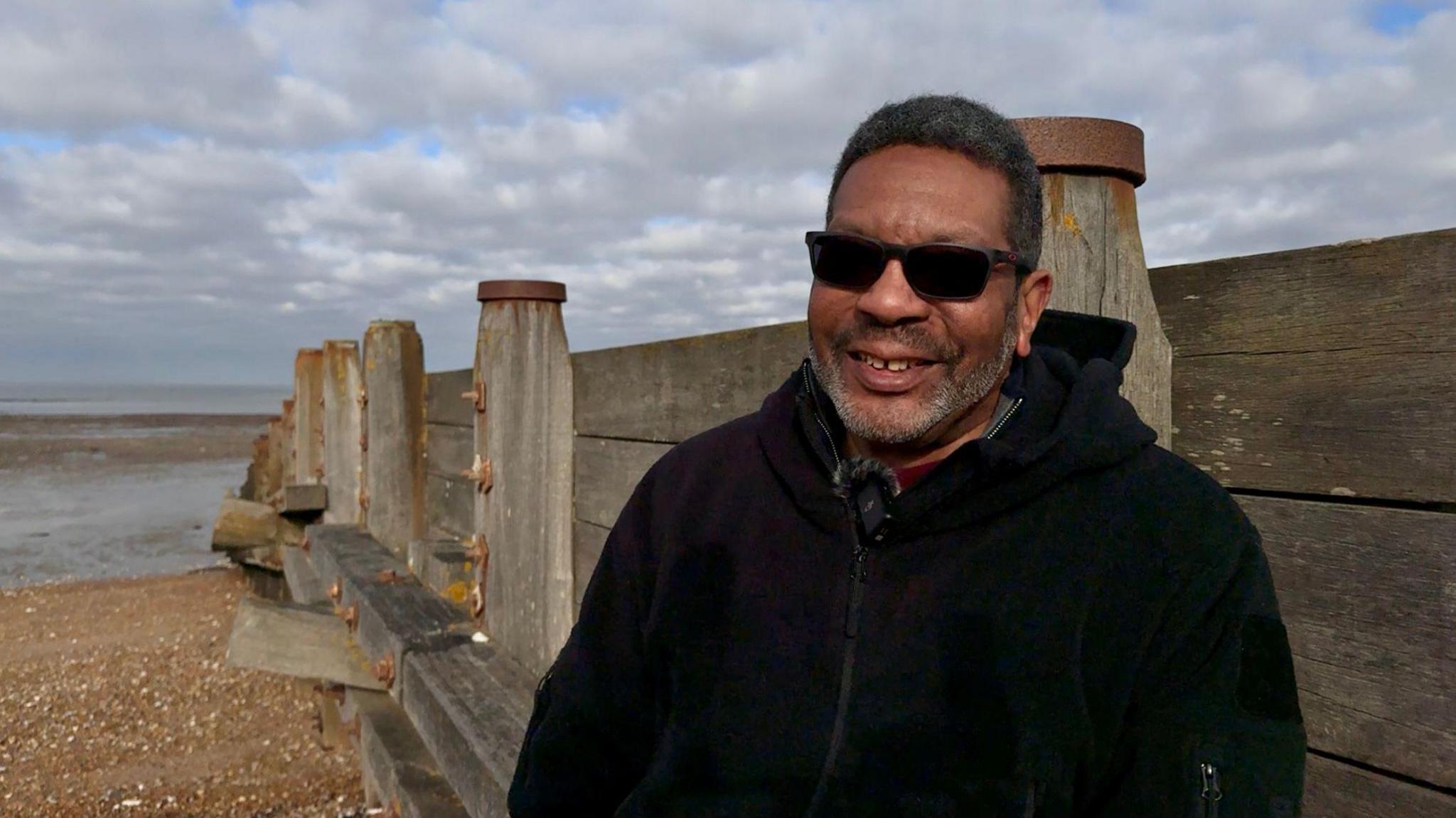 Fred Jackson stands on a beach in front of a large wooden structure and with the sea visible in the background. He is wearing dark sunglasses and a black zip up hoodie.