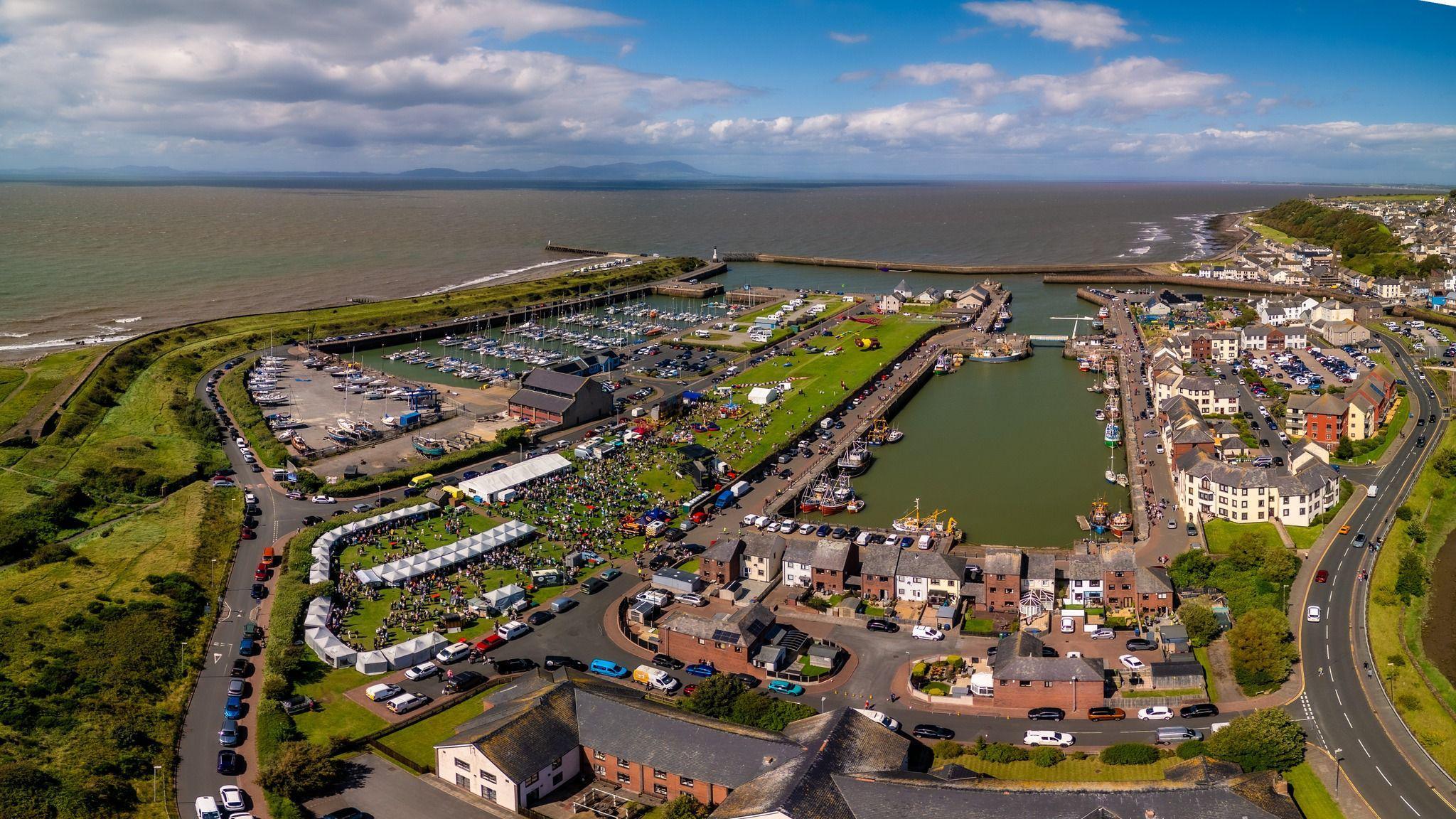 Aerial image of Maryport's harbour area. A crowd of people can be seen around an area with several stalls and music stage.