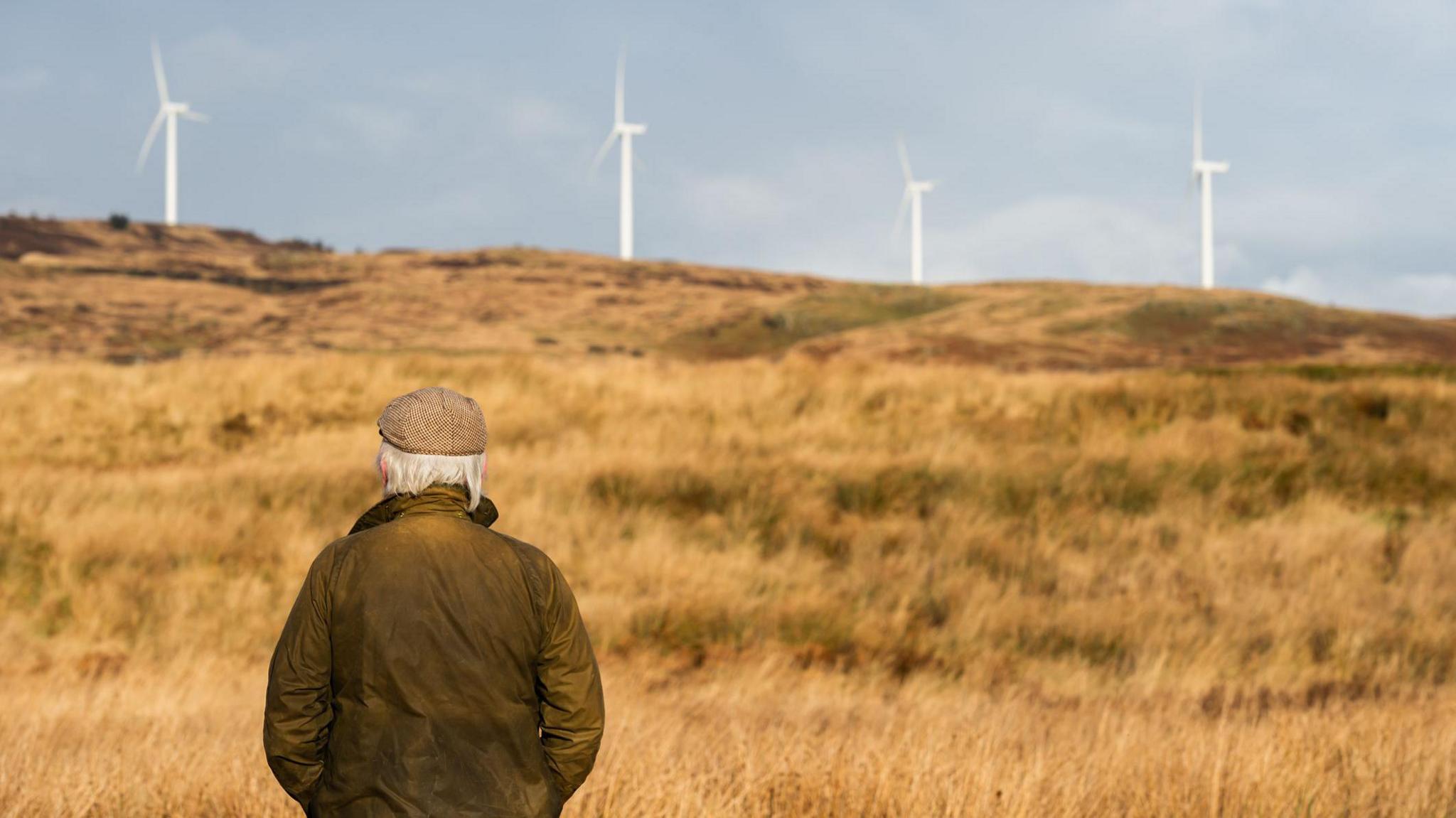 A man in a jacket and bunnet stares out across grassland towards four wind turbines in the distance