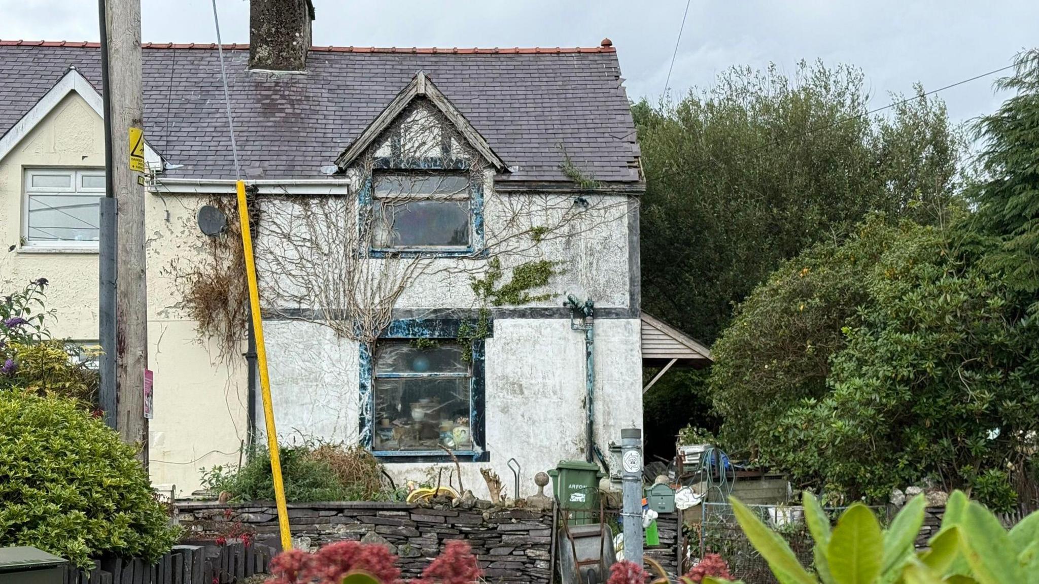 A wide image of the exterior of the detached white house. It is a two-storey house with a front and side garden. The house has stains on and Ivy covers the upstairs. 