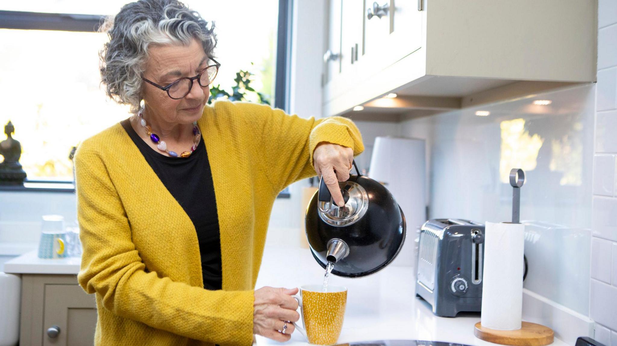 Older woman in a kitchen pours hot water from a kettle into a mug.