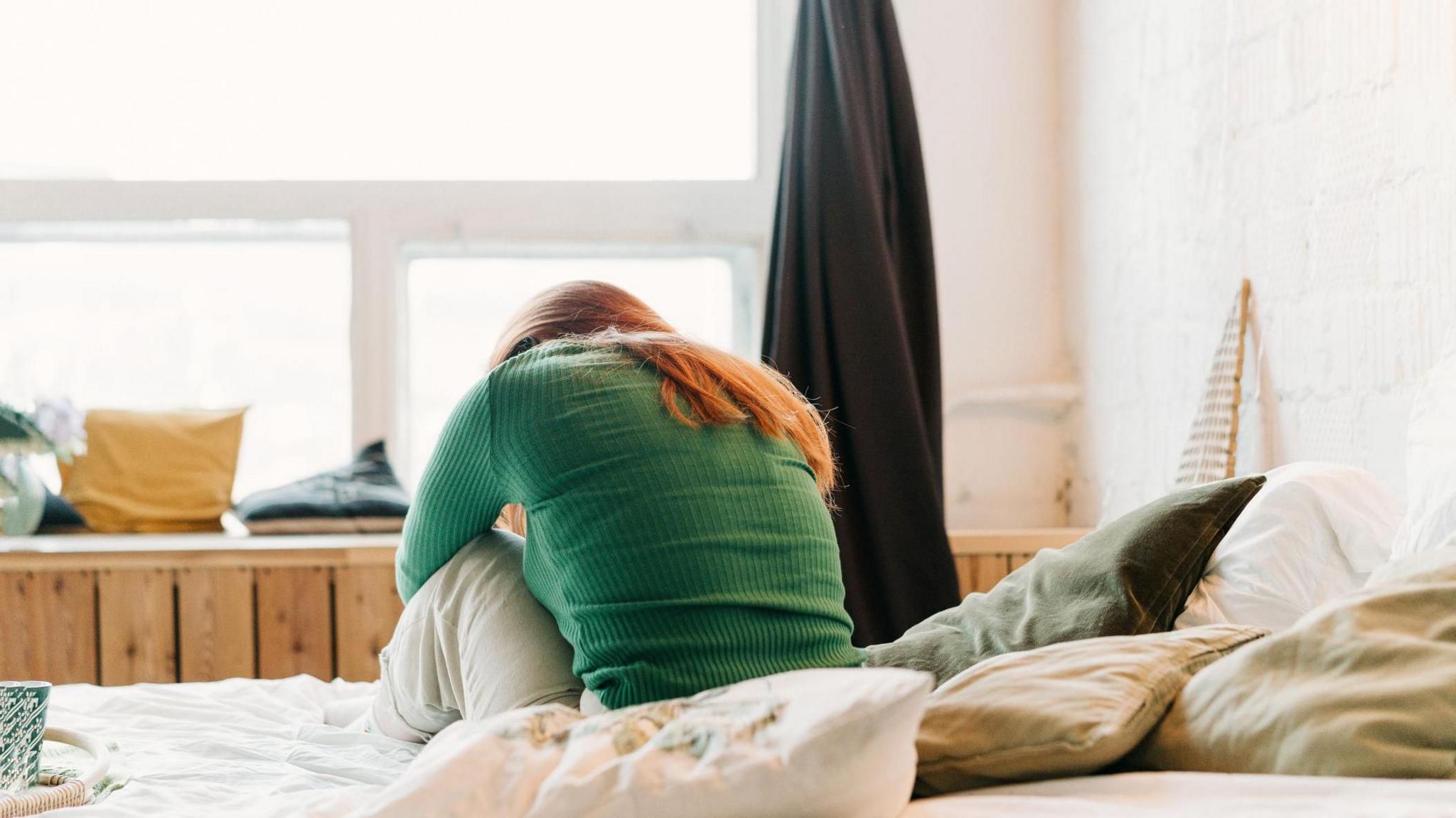 A young person is sitting on a bed, they have their back to the camera and have long dark hair. There are cushions on the bed.