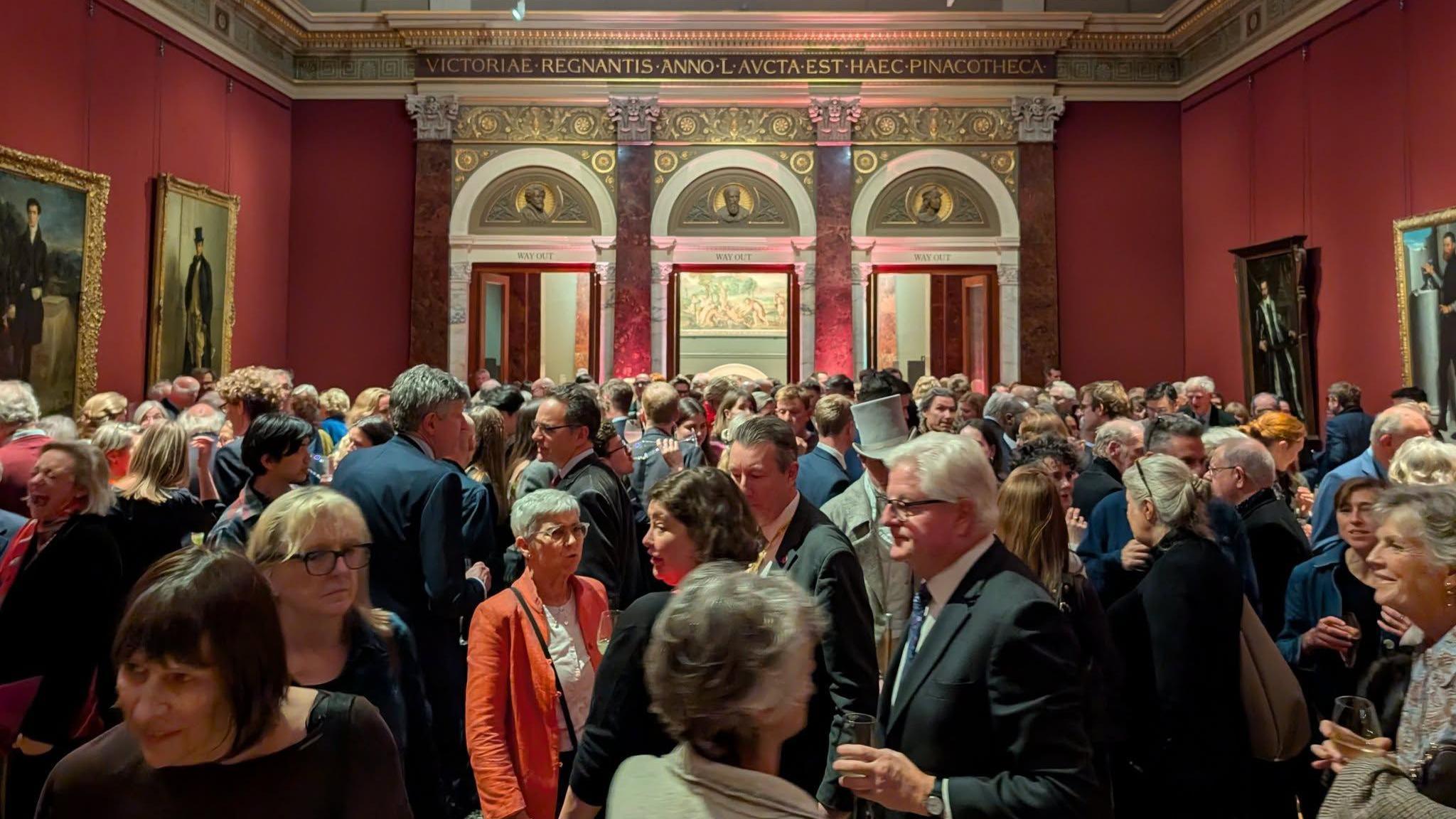 Crowd of people looking at paintings in a large red room