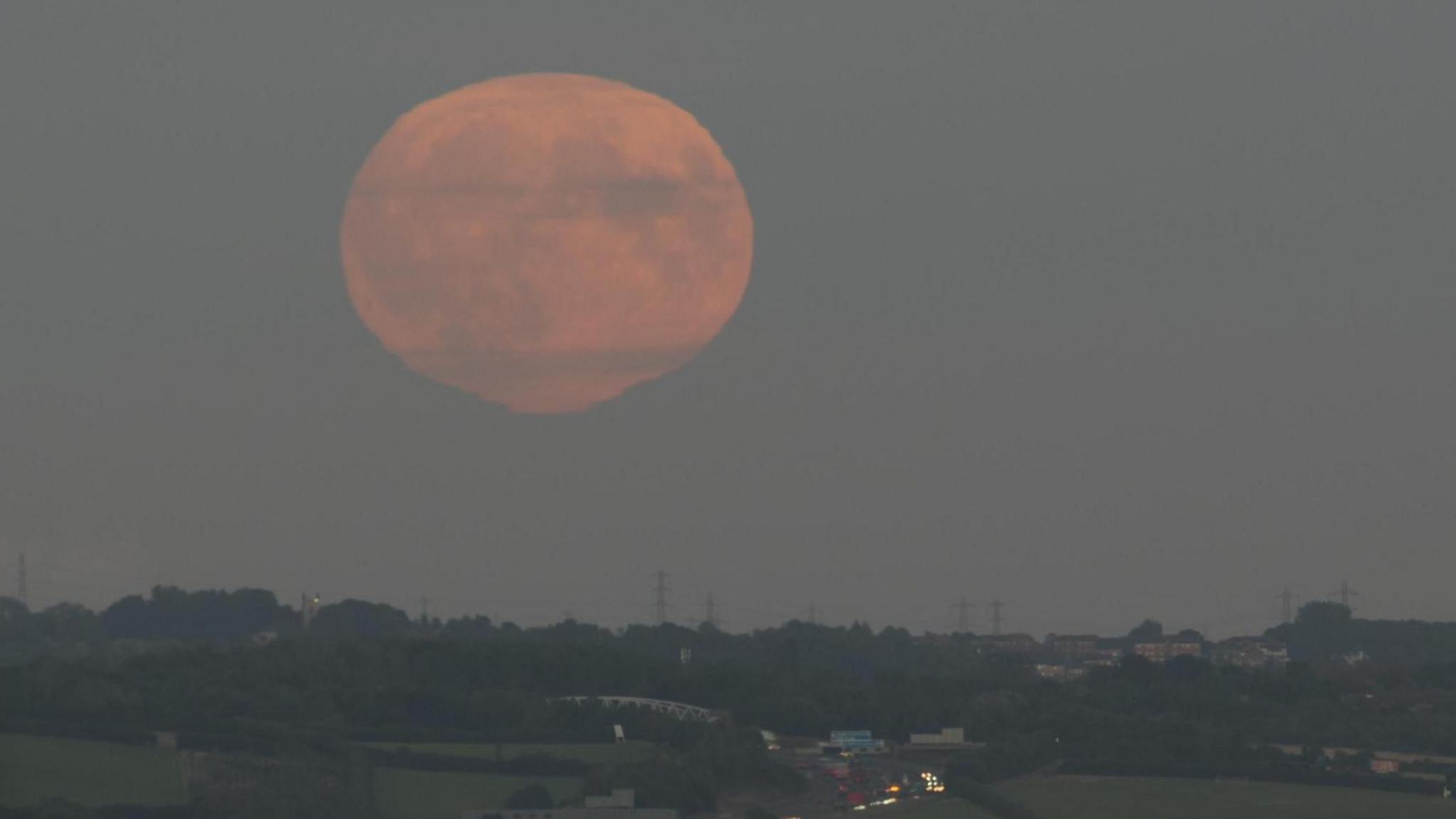 A coppery supermoon above Armley in Leeds. The city's skyline can be seen in the distance behind green hills and trees.