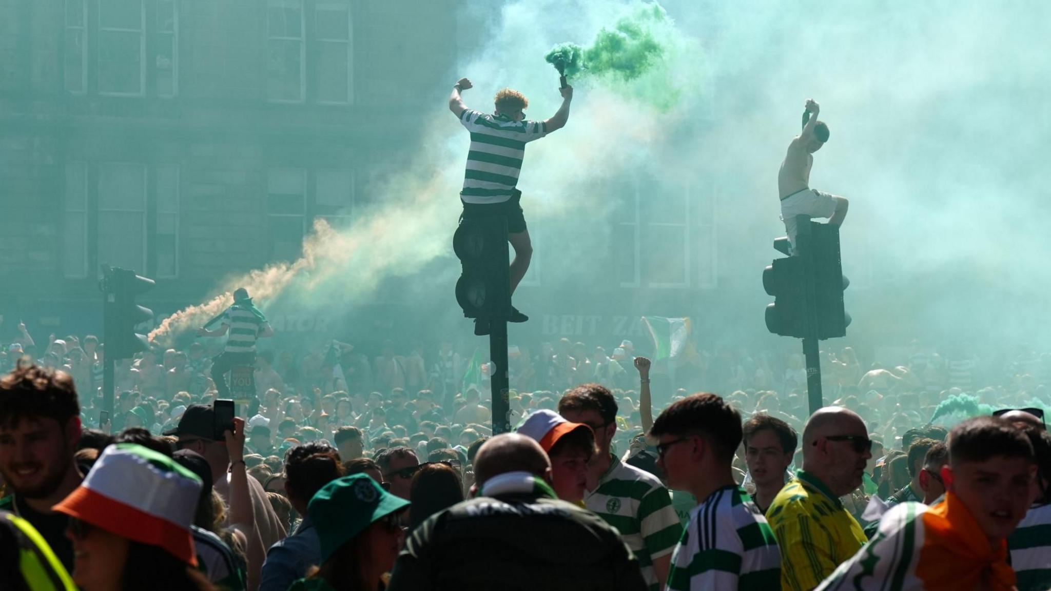 Celtic fans sitting top of traffic lights with hundreds of other fans on the street below them. One fan on a traffic light is holding a smoke bomb and there is more smoke at the back of the photo