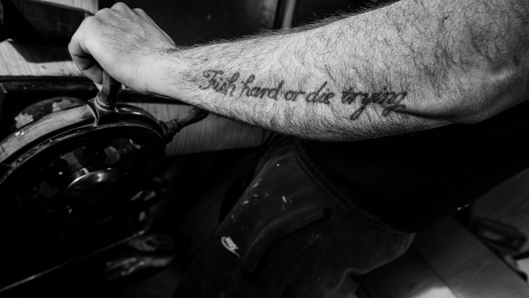 A black and white image of a fisherman's arm at the helm of a boat. A tattoo in cursive writing reads ' Fish hard or die trying'. 