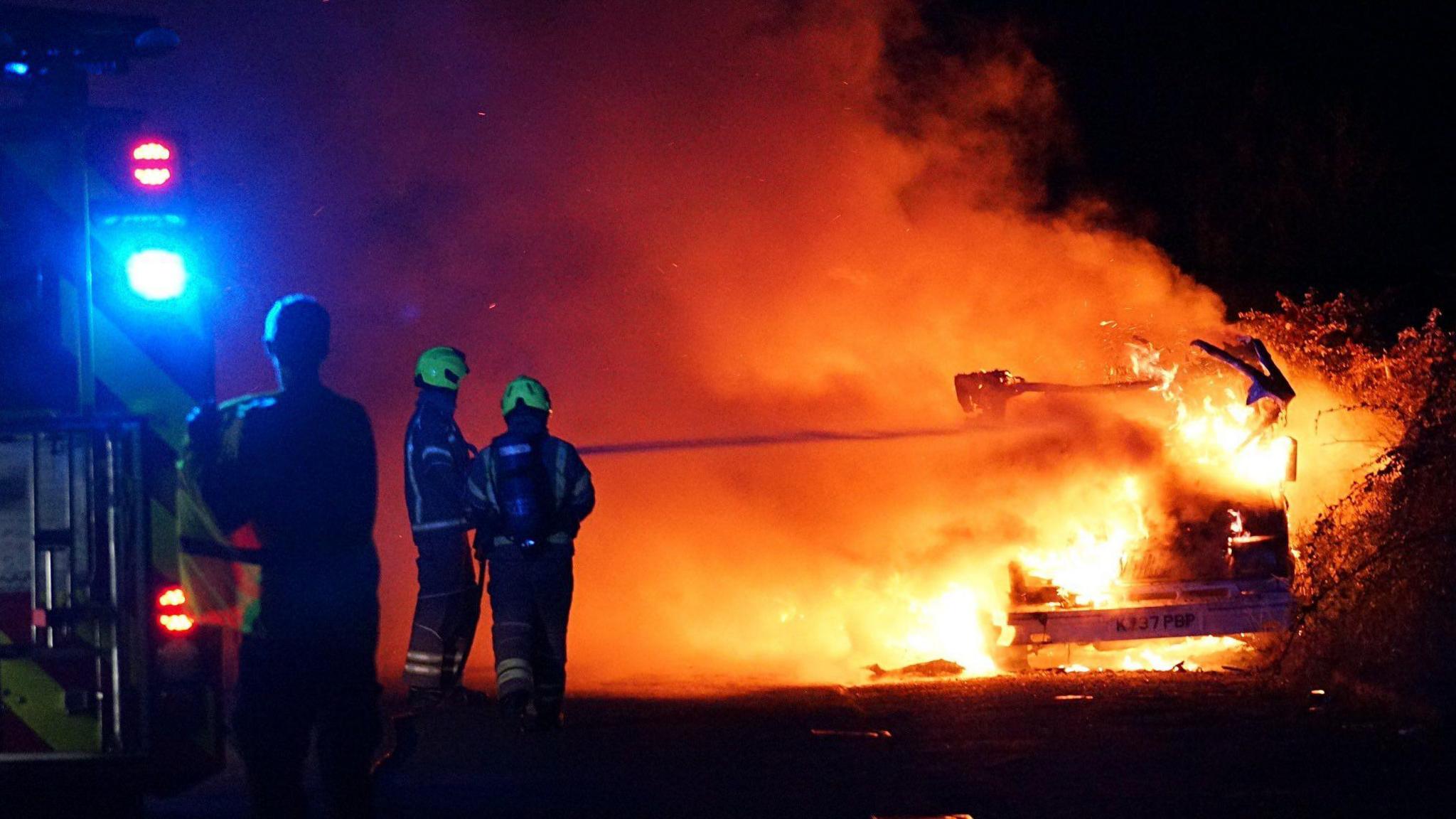 Two firefighters firing a hose of water into a camper van which is on fire. It is dark and there is a fire engine on the left