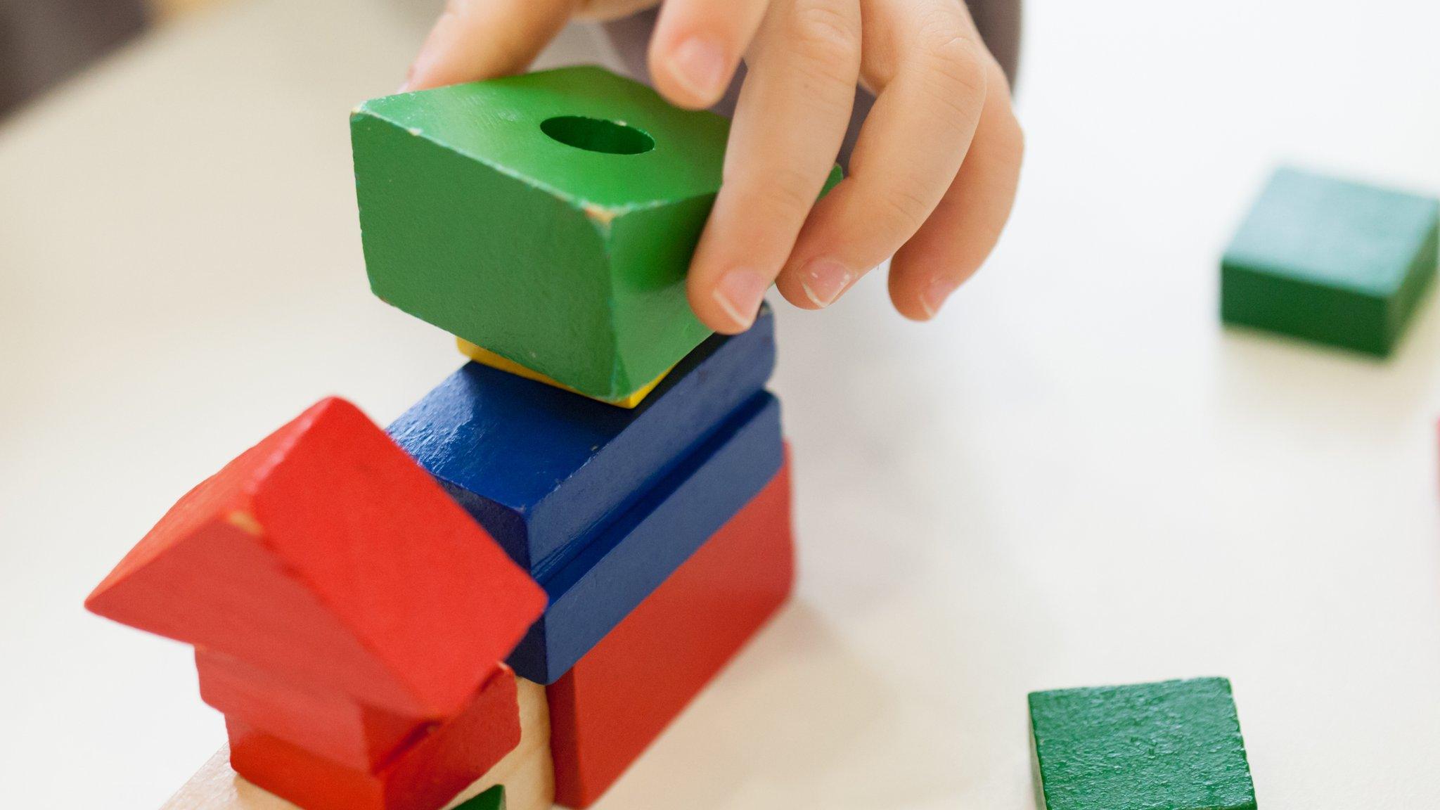 Child's hand playing with coloured wooden brick shapes on white table