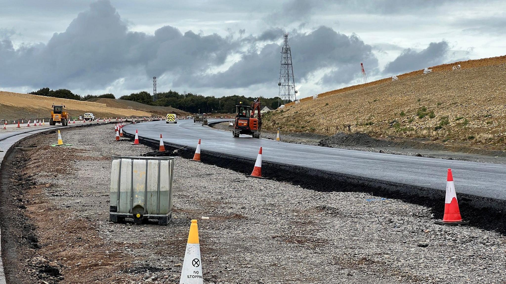 A freshly tarmacked road on a large building site in the middle of nowhere. There are large mounds of dirt and earth on either side and traffic cones, with construction vehicles driving along the road.