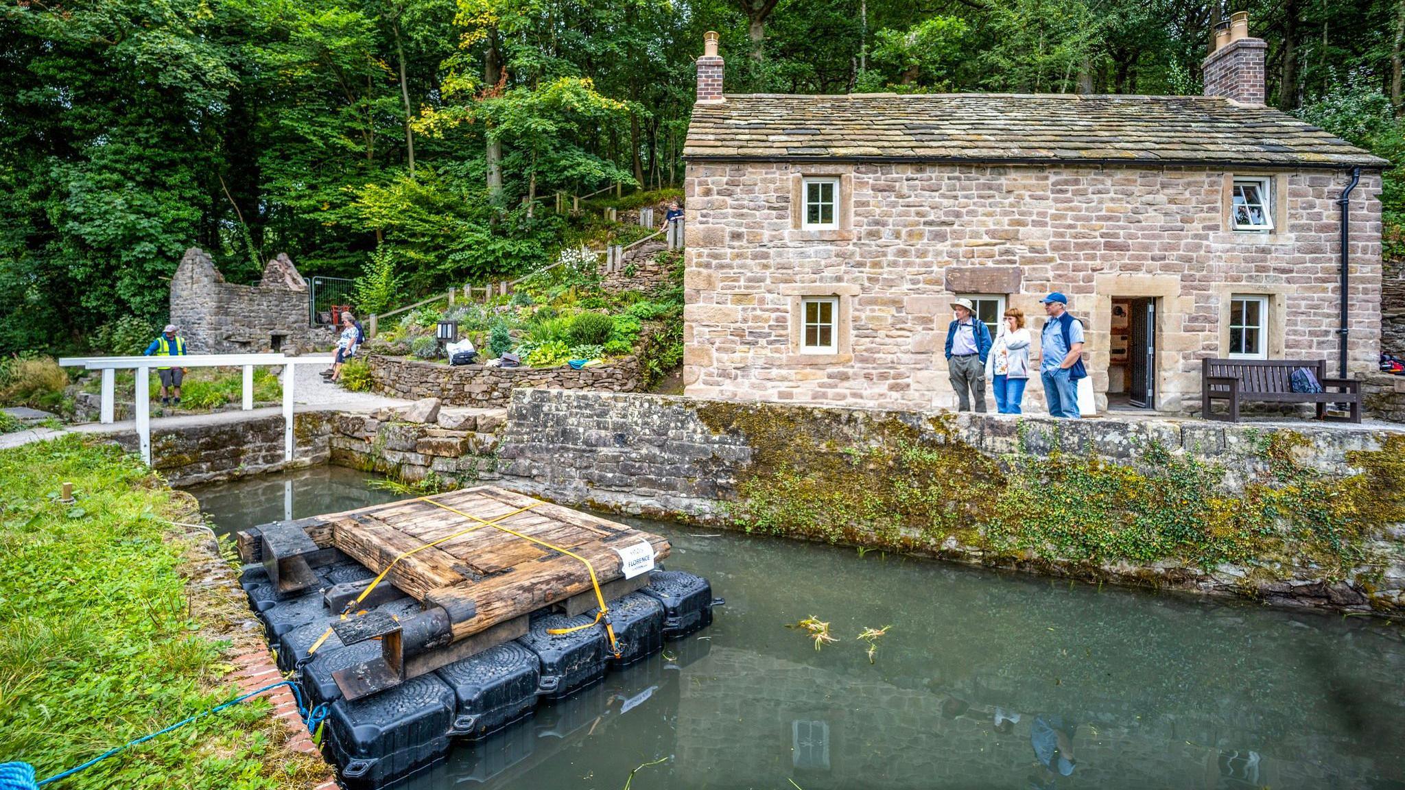 Aqueduct Cottage, a Grade II listed 19th Century lock keeper's cottage, which has light coloured stone work. The cottage has a wall in front of it before a canal. On the canal is the lock bridge on a pontoon