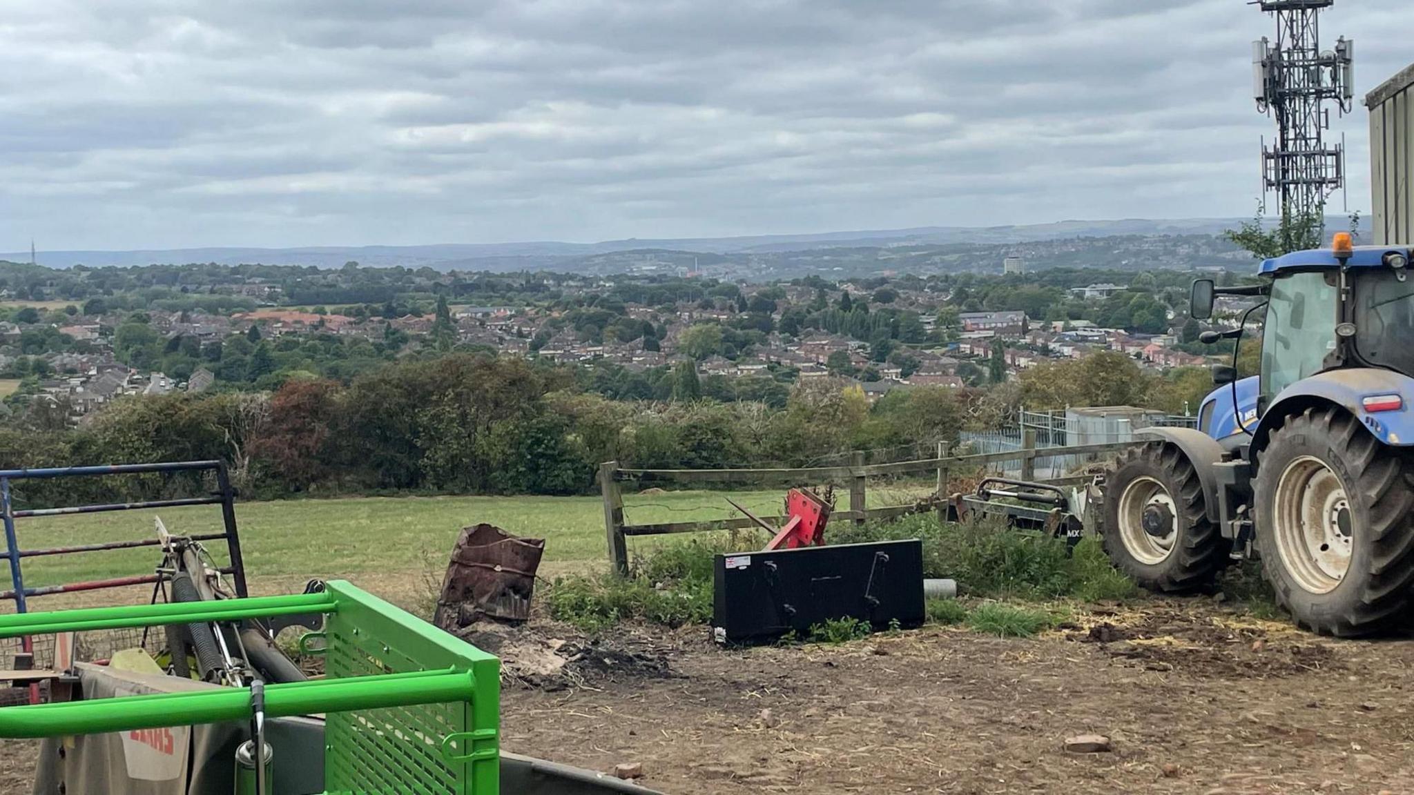 A muddy farm with a blue tractor and green machinery overlooking an urban area with houses.