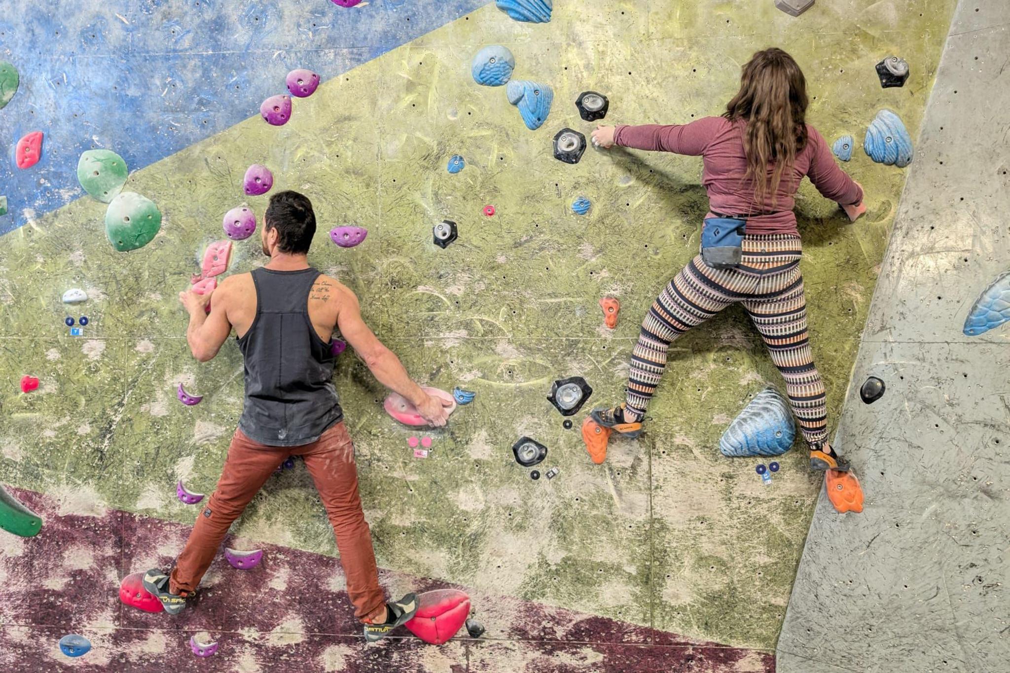 A man and a woman are climbing on a multi-colour climbing wall. They are both facing away from the camera