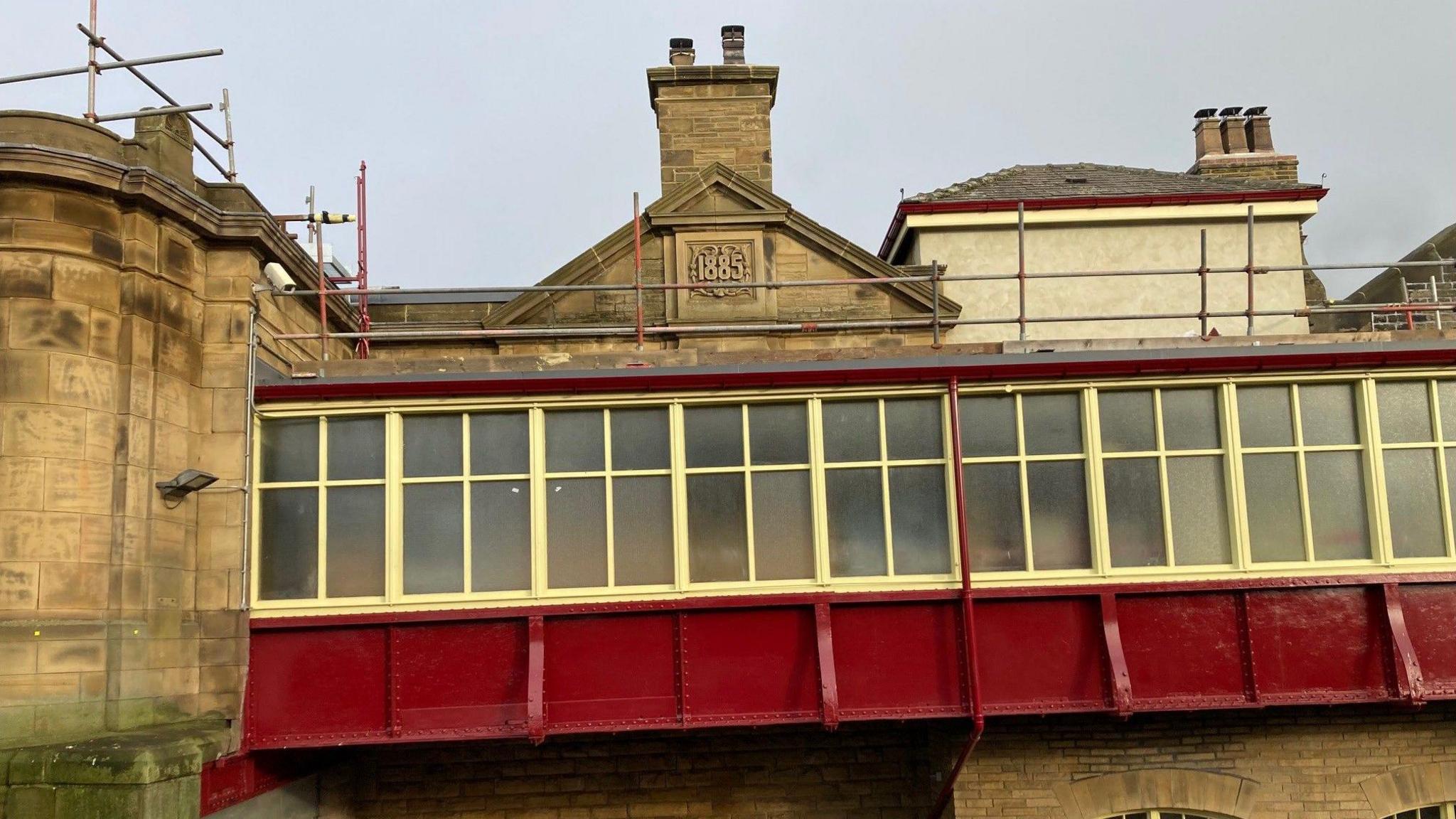 The outside of a Yorkshire stone building with a metal walkway painted in maroon and yellow. A date stone on a chimney stack above reads 1885.