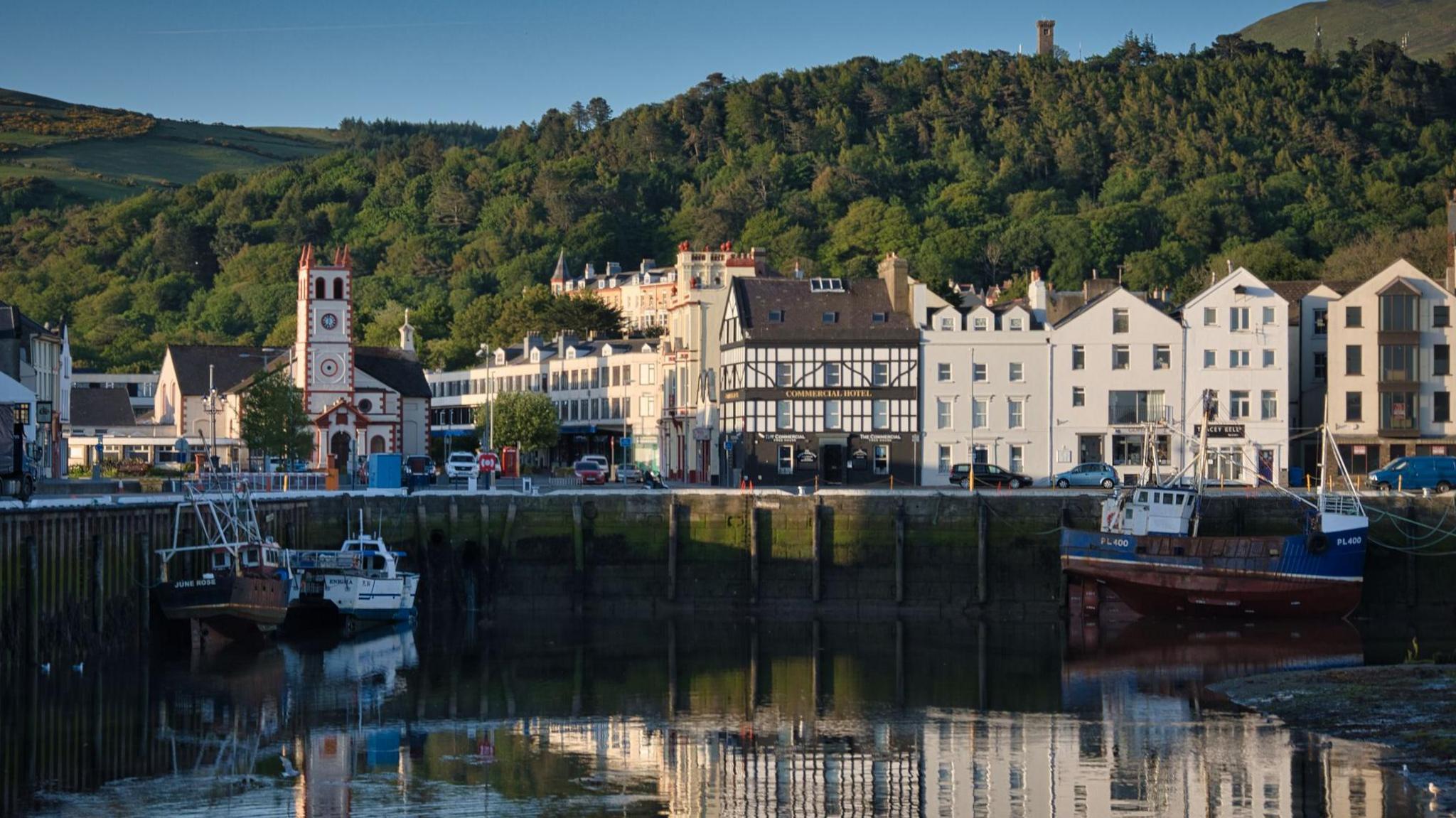 A view across Ramsey harbour at low tide. There are fishing boats low in the water against the quayside lined with townhouses and a church on the left. Behind is a green tree-filled hillside with a small tower at the summit to the right.
