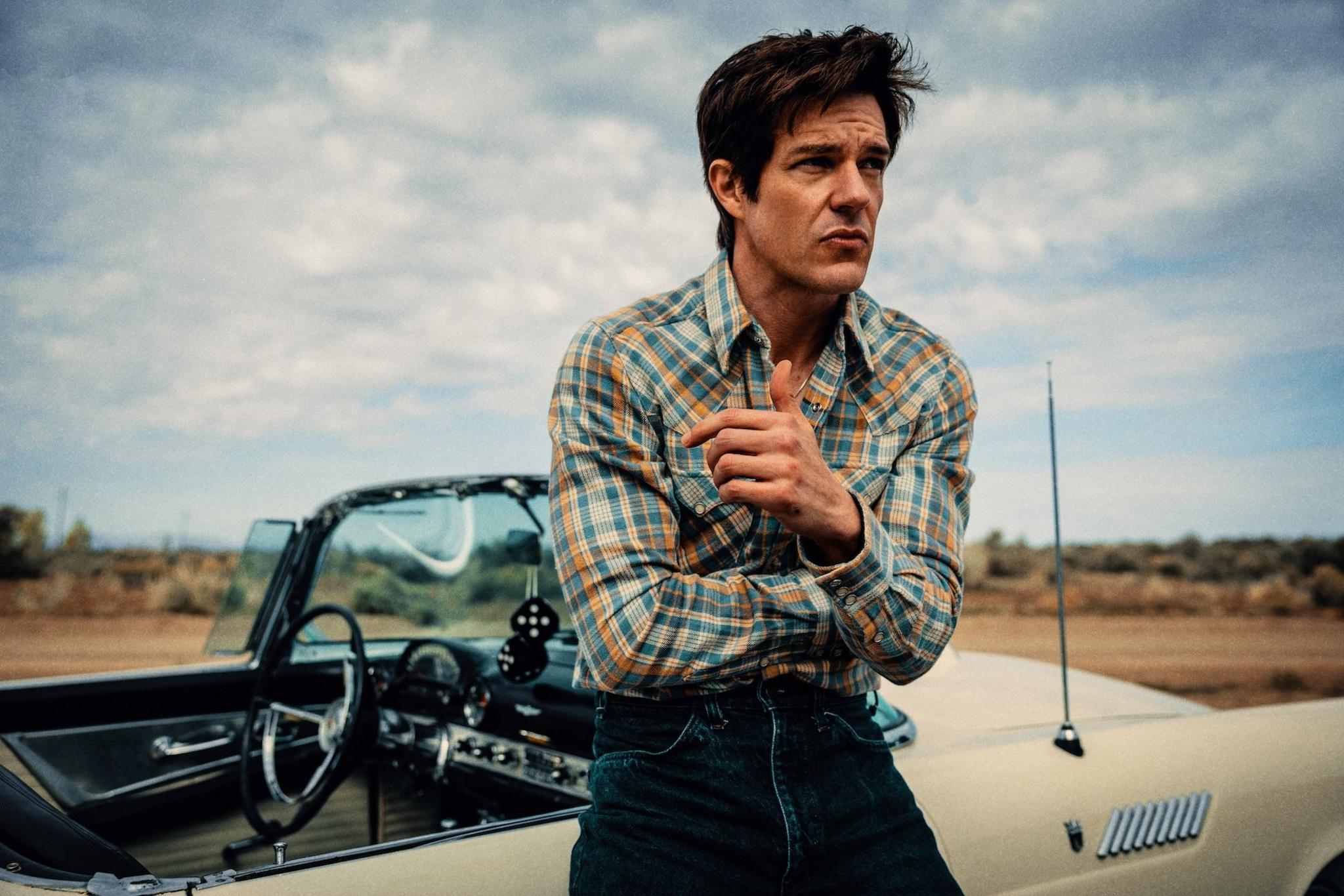 Brandon Flowers in a checked shirt, leaning against a classic American convertible, with fluffy dice hanging from the rear view mirror