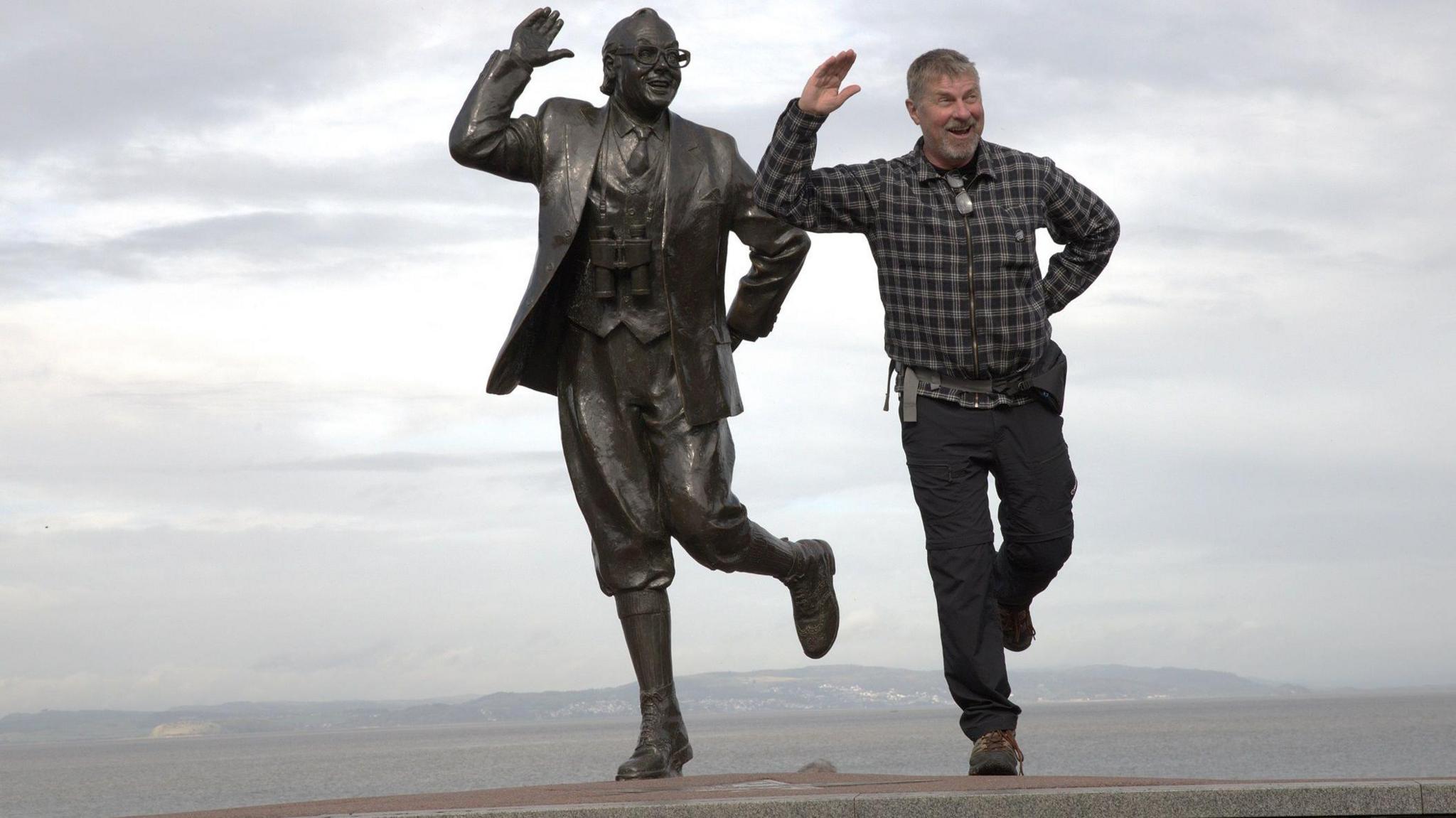A man wearing a checked shirt and black trousers posing next to a statue in Lancashire.
