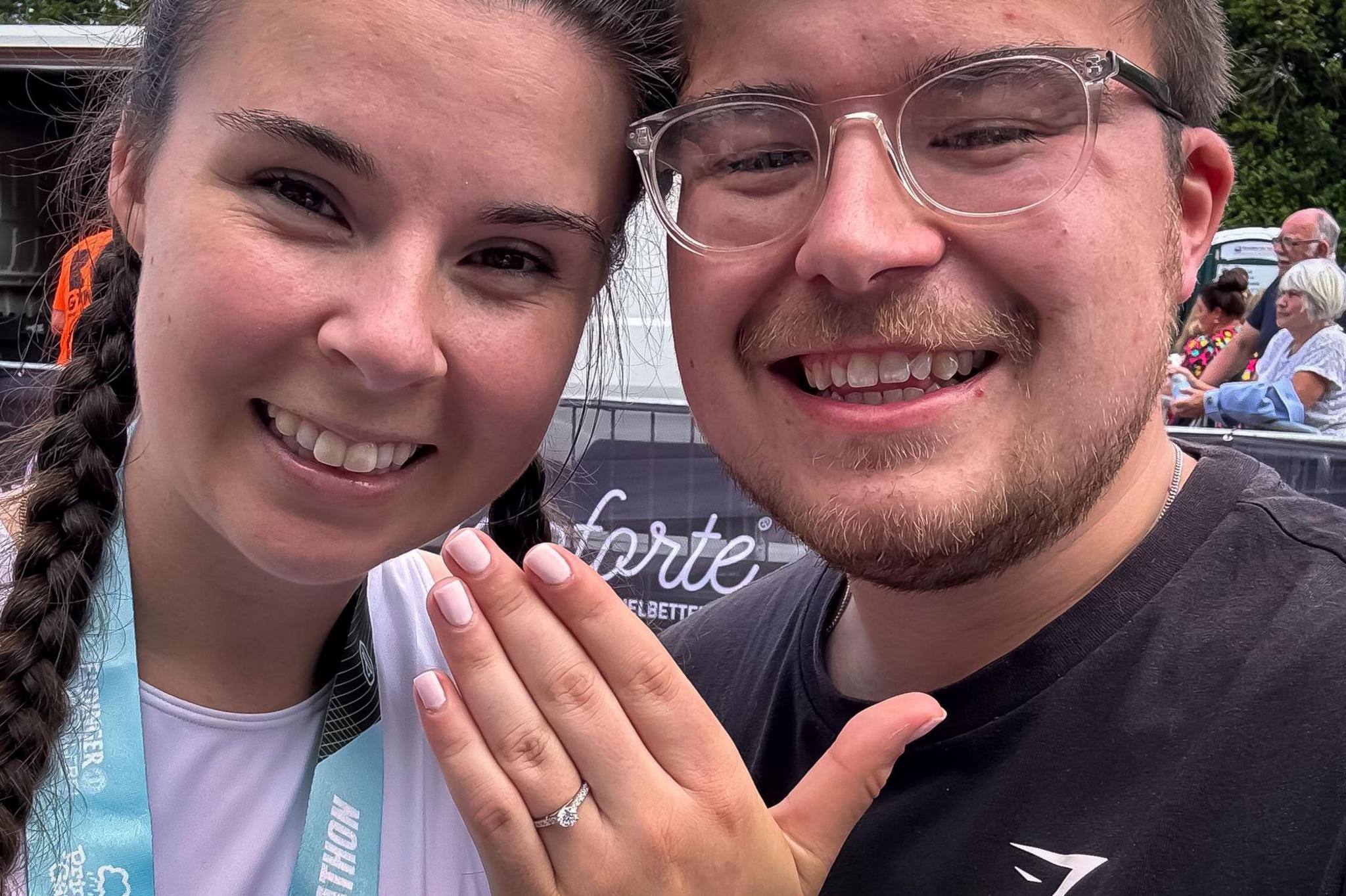 Sarah holding her hand up with an engagement ring beside Thomas, both smiling