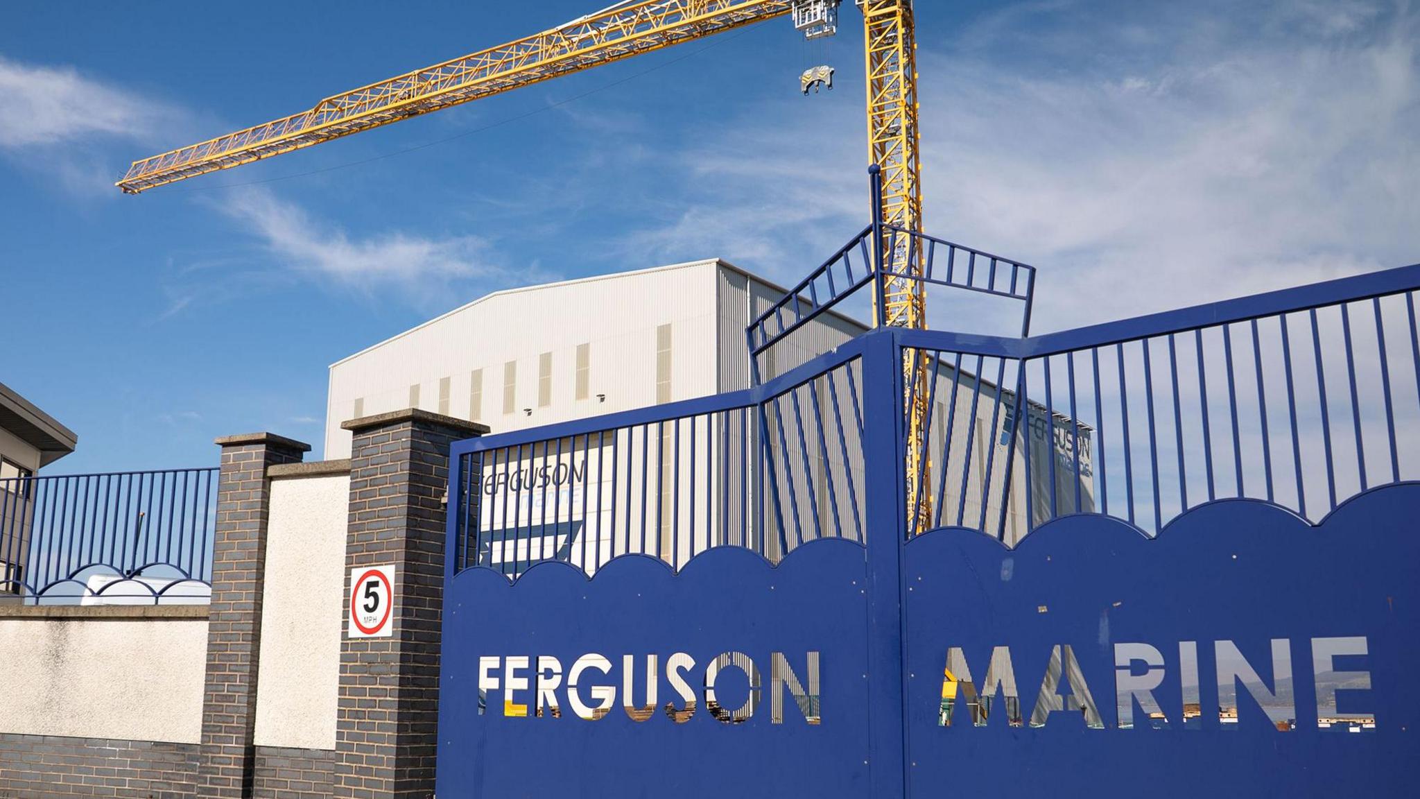 A blue gate with the words Ferguson Marine cut out. A large module hall and yellow crane can bee seen in the background