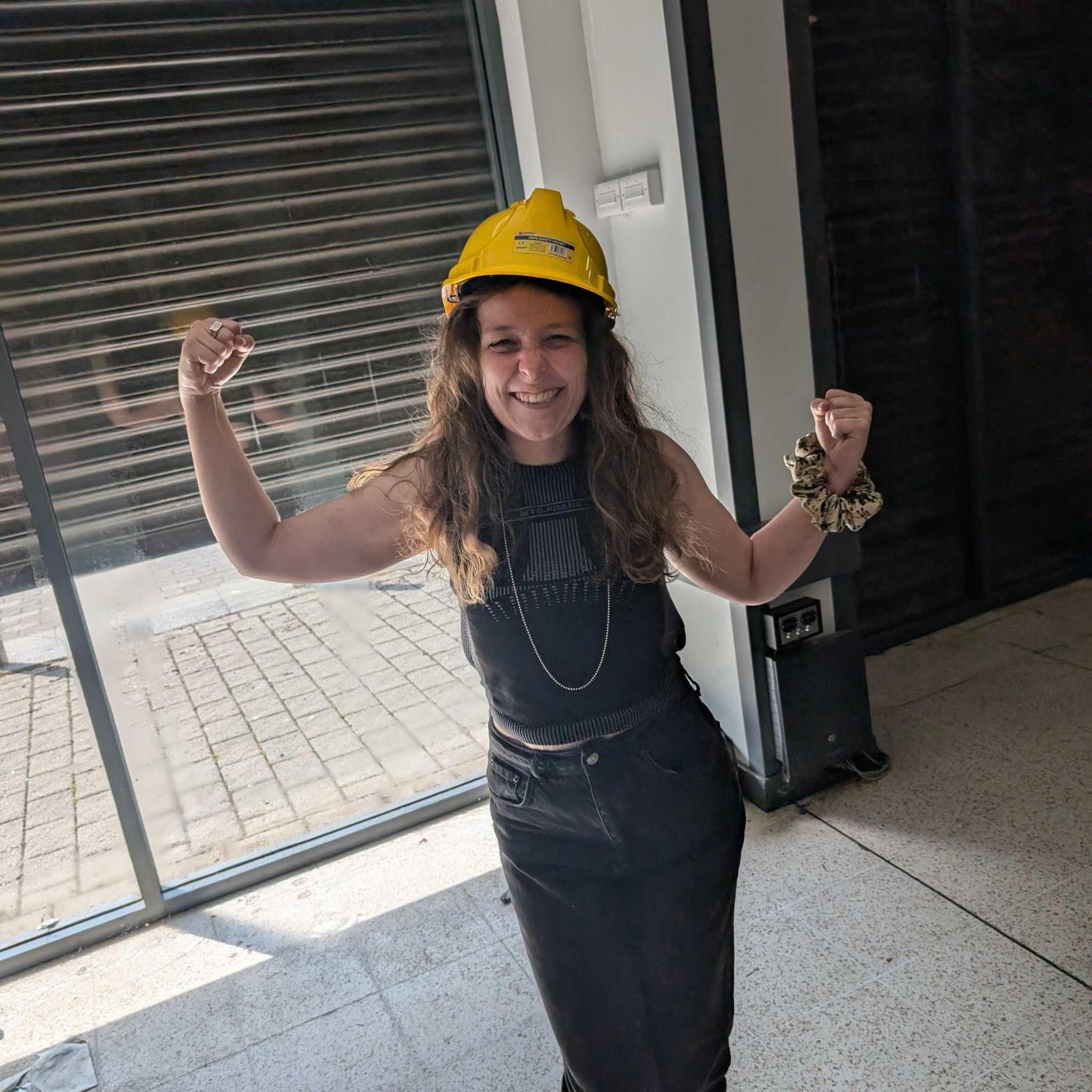 Laura Robertson standing in the empty store in front of its doors. She is wearing a yellow hard hat and is flexing her biceps in a celebratory strongwoman pose. She has long brown hair which hangs over her dark clothes.