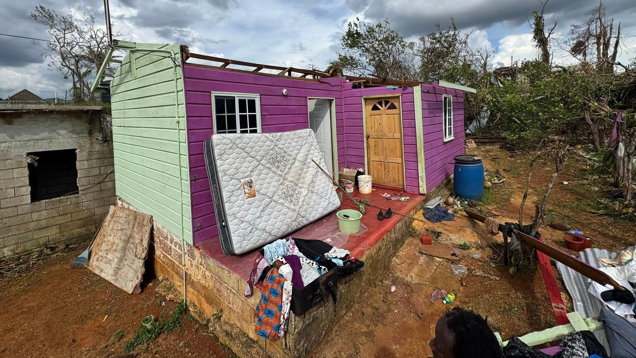 A house made of wood painted purple and green with a wooden door. There is a small brick building behind it. There is a mattress in front of the house and debris on the ground. The building has no rood. There are fallen trees. The sky is grey and cloudy. 