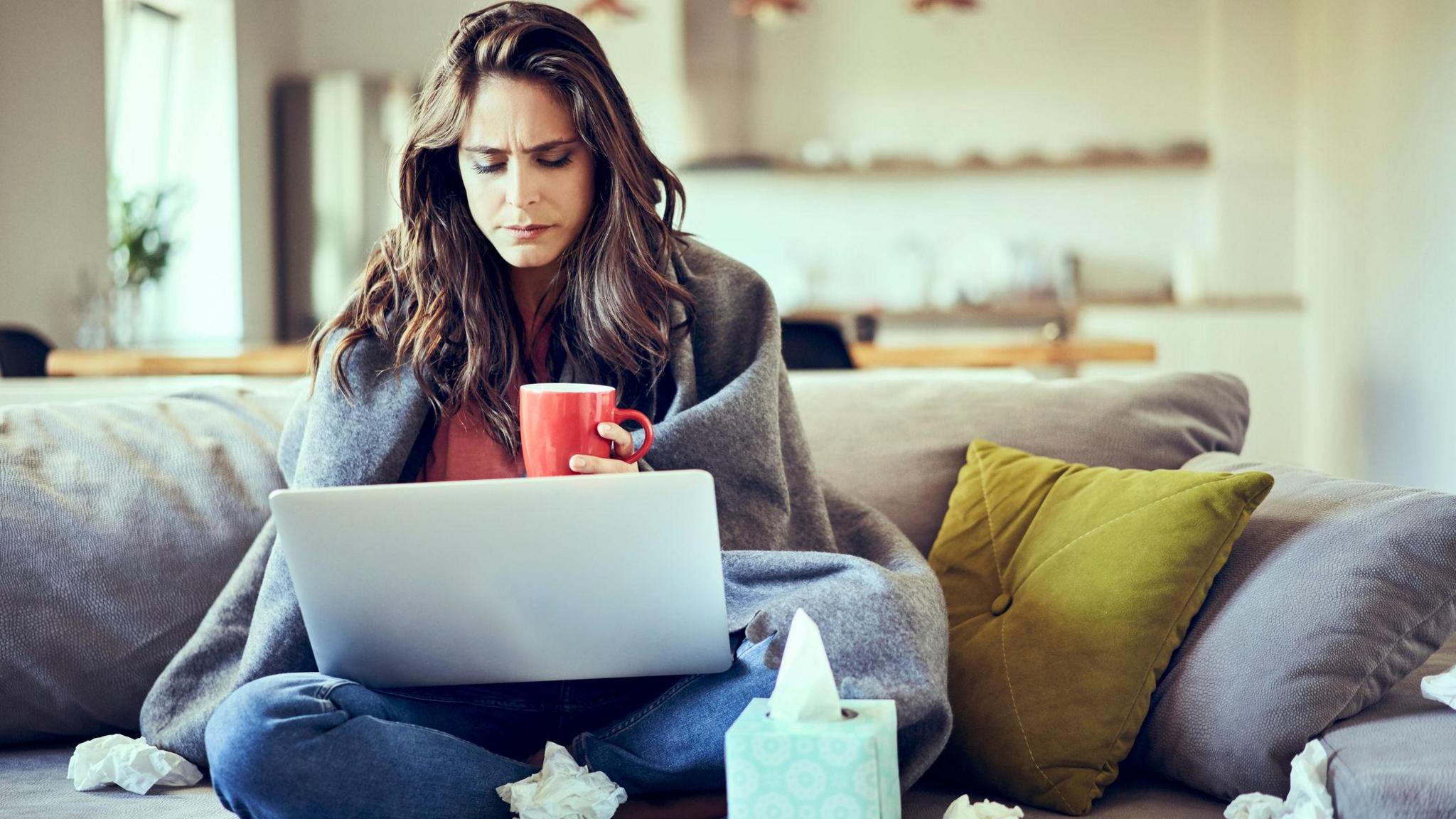 A sick worker sits at home on their laptop wrapped in a blanket surrounded by used tissues