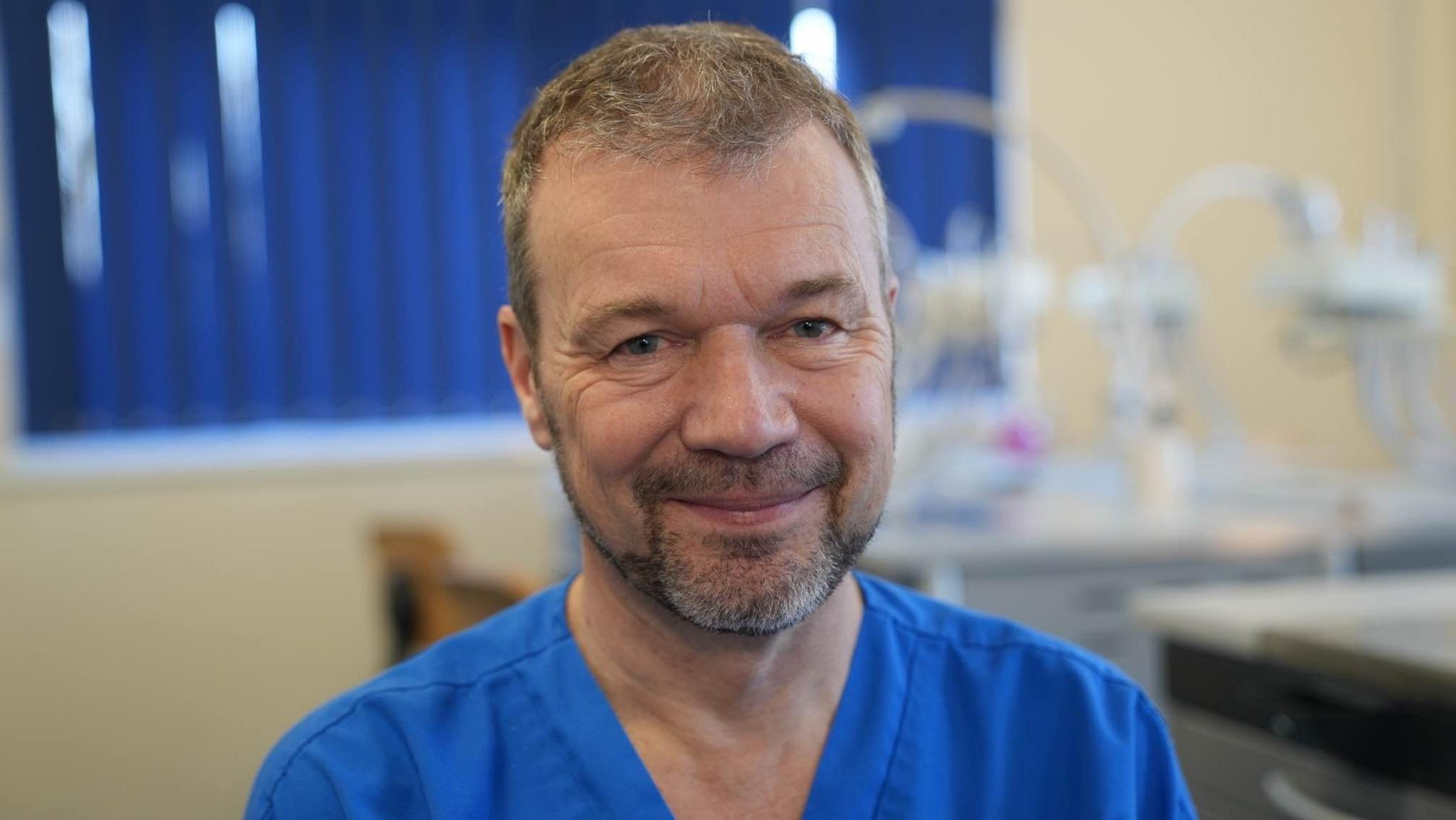 Professor Nicholas Barker is wearing blue scrubs and looking at the lens of the camera, whilst smiling. The background which is of a dentistry training room is slightly blurred and machinery can be seen over his left shoulder.