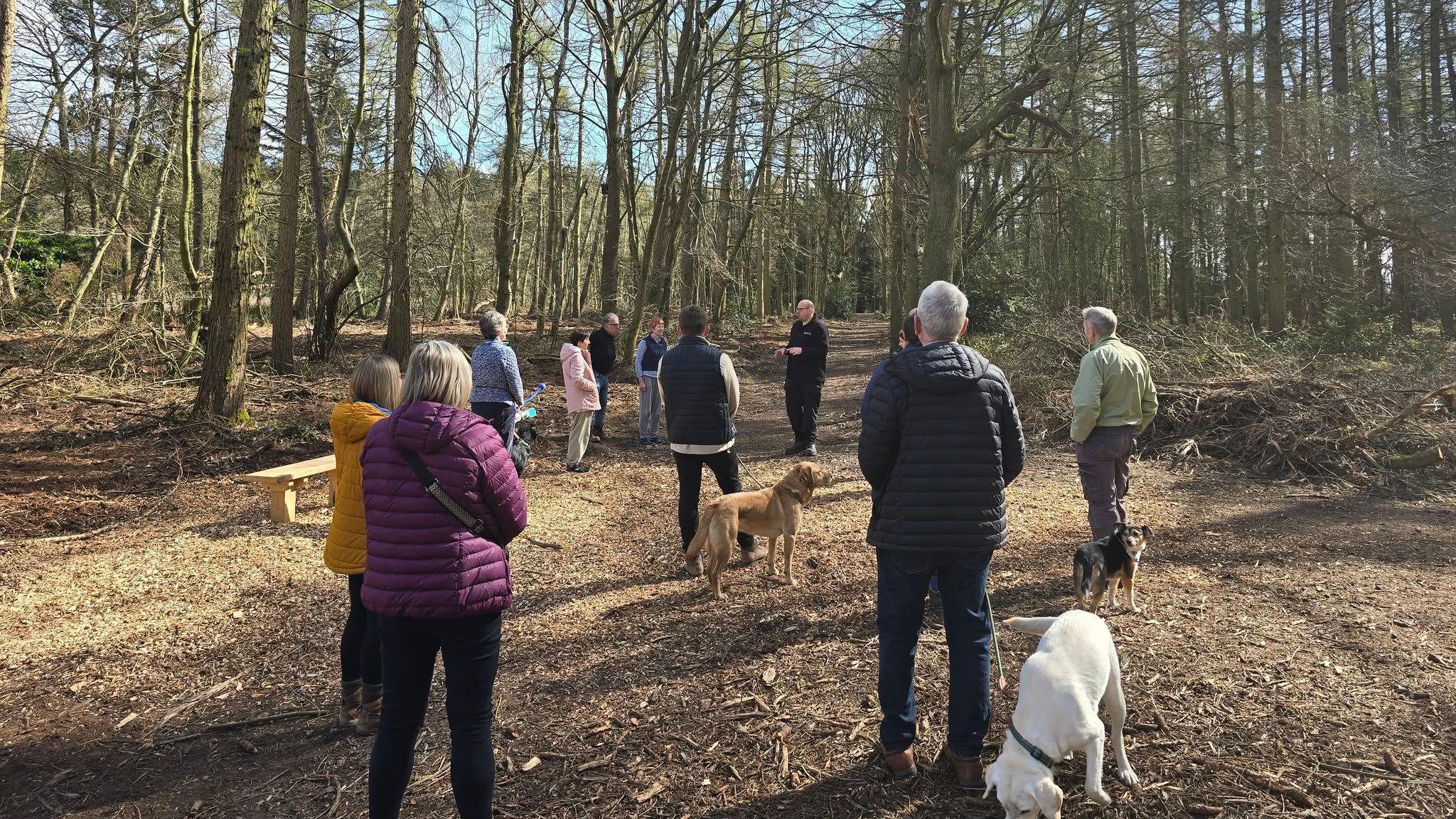 A group of dog walkers wearing coats and with dogs on leads stand on a path in a wooded area.