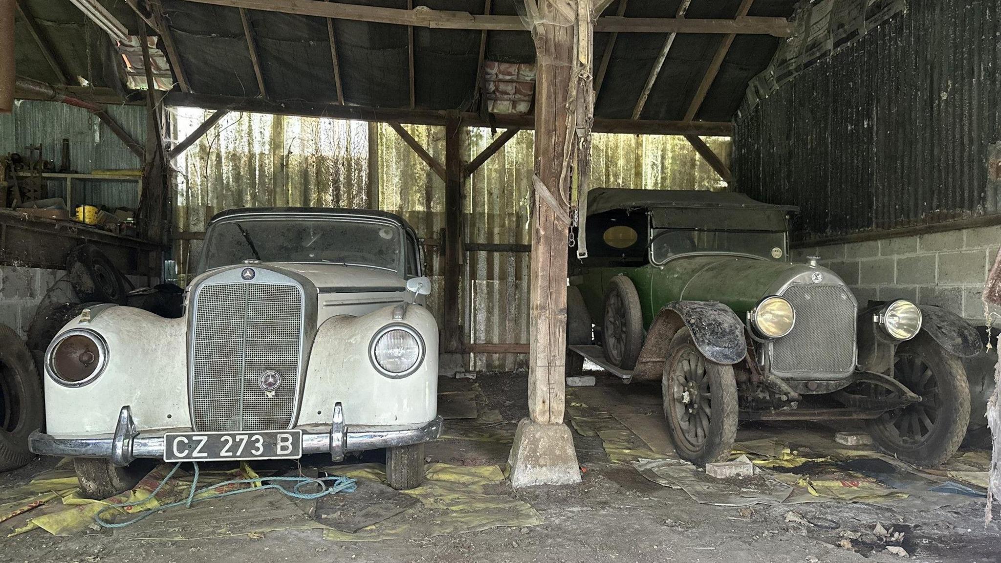 Two dusty cars - a white Mercedes from the 1950s and a 1921 green Talbot - pictured with bonnets facing the camera in a dilapidated barn