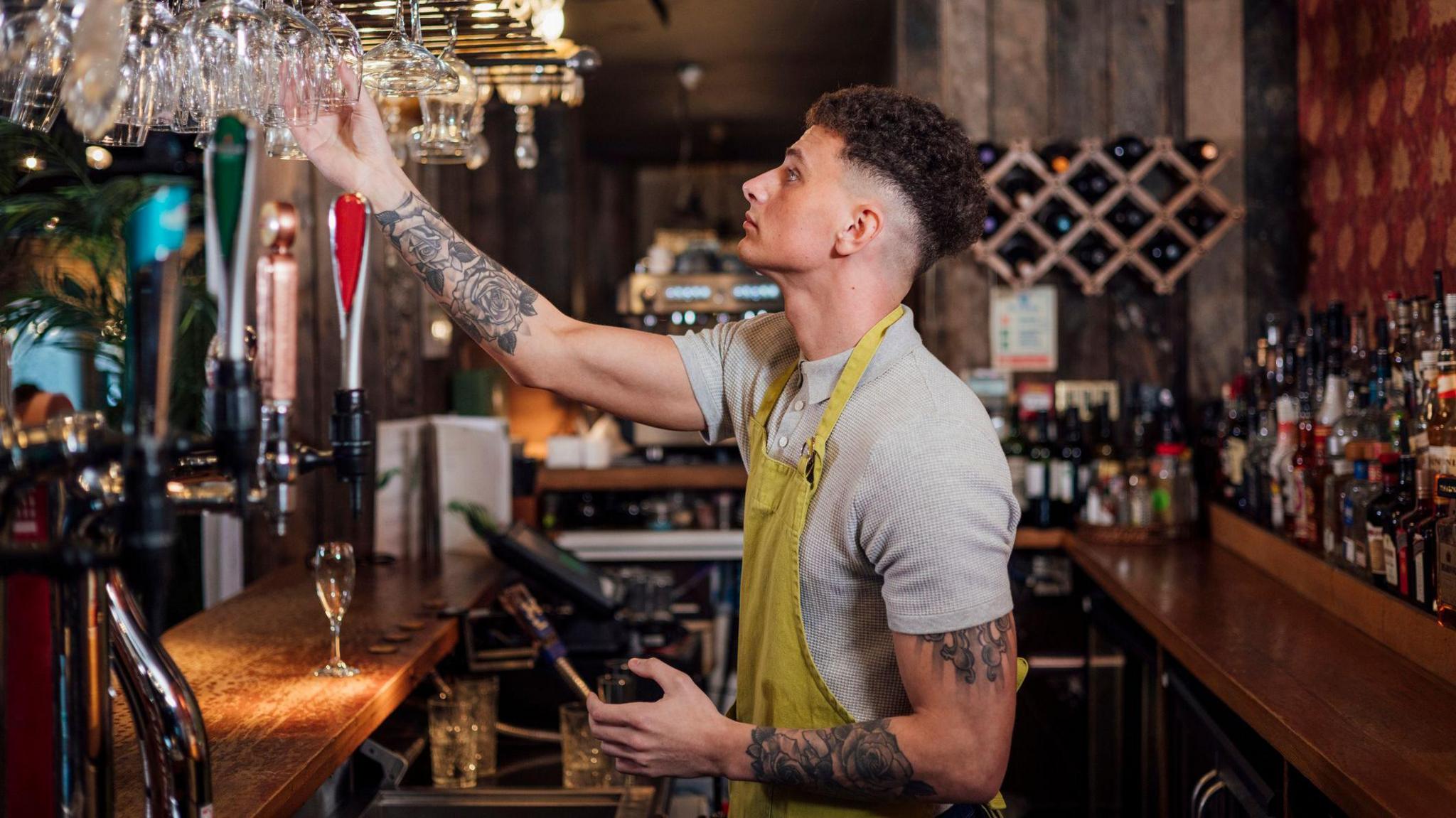 A bartender wearing a grey polo shirt with a yellow apron on top hangs up glasses after washing them.