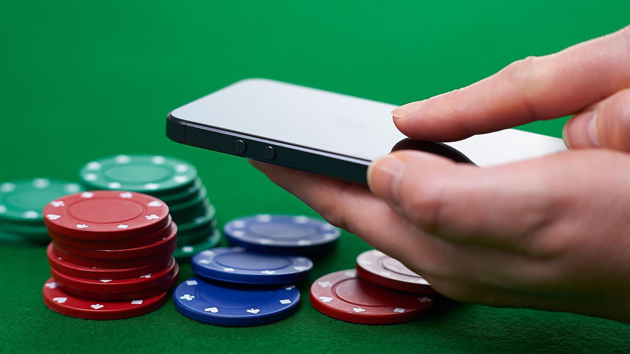 A hand holding a black smartphone, hovering over a selection of green, red and blue poker chips