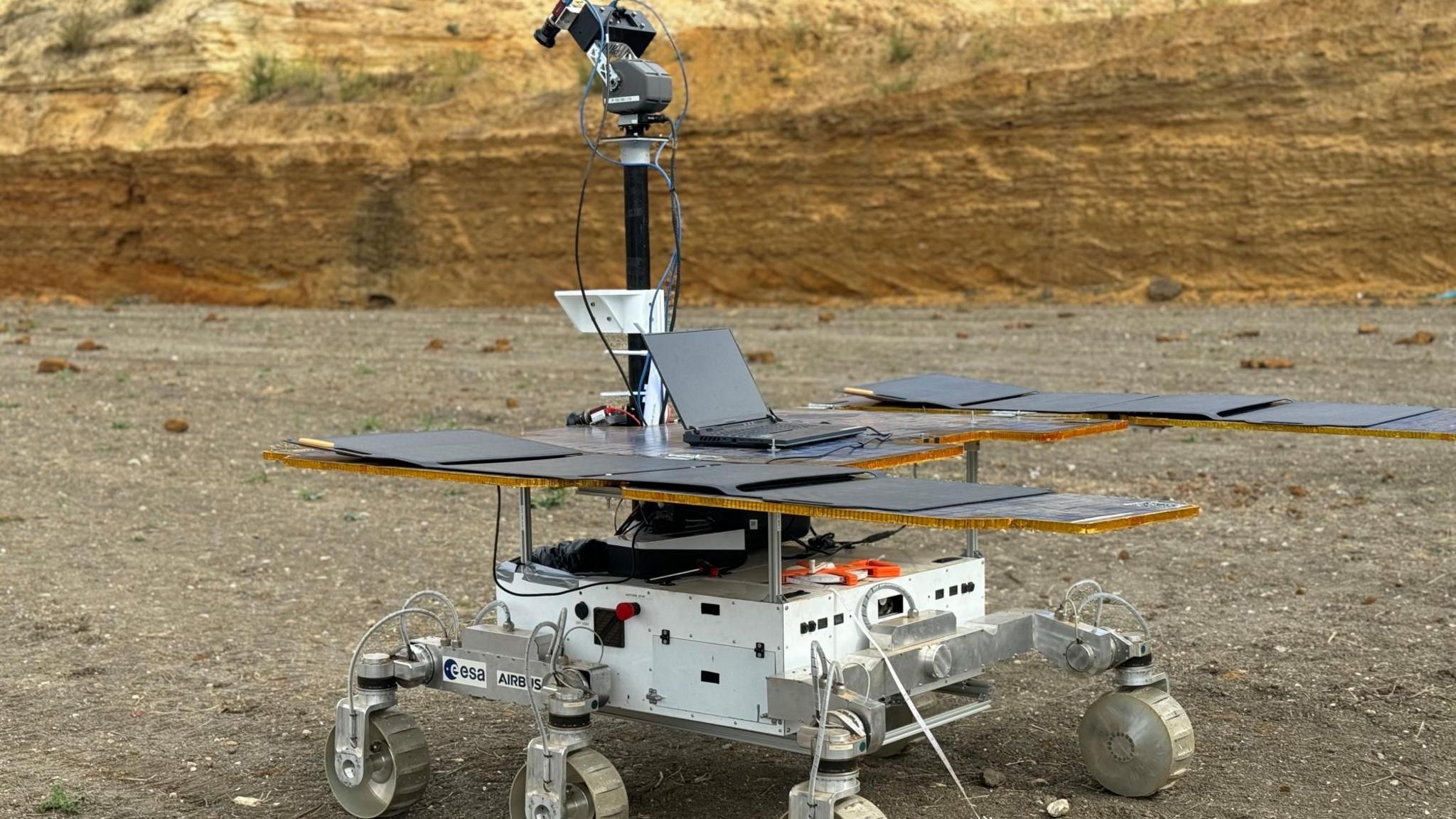 A six wheeled Mars rover is parked in a quarry, it had large solar panels and a camera fixed on top. A laptop is resting on top of the rover