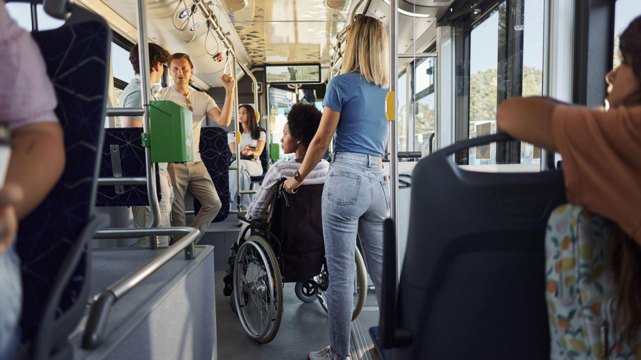 A woman sitting in a wheelchair on a bus, with another woman standing behind her holding the wheelchair handles. There are five other people on the bus.