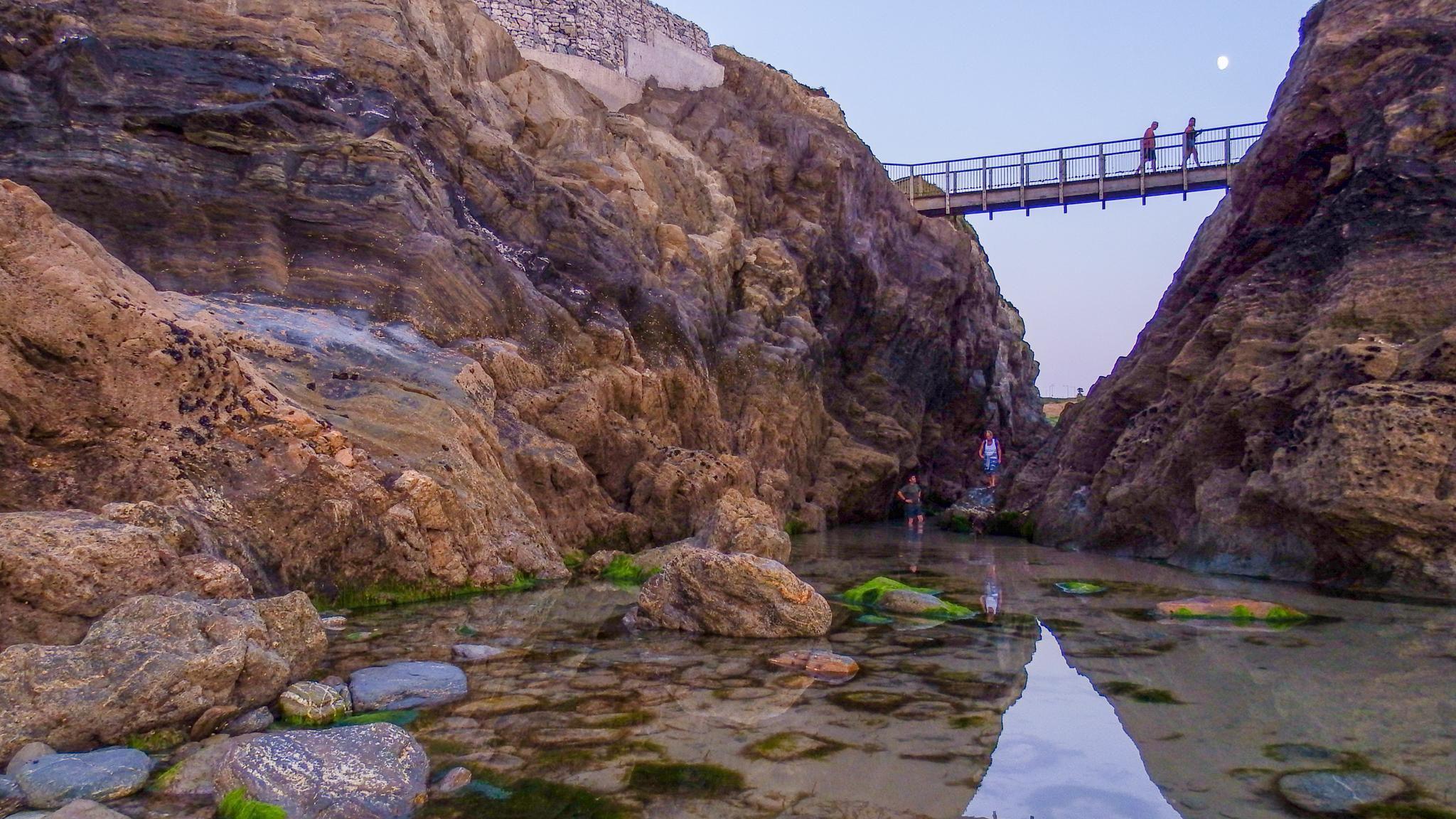 People look around part of Porth in Cornwall. The area is a large rock pool. Two people are stood near the water. Two other people are walking along a bridge above them.