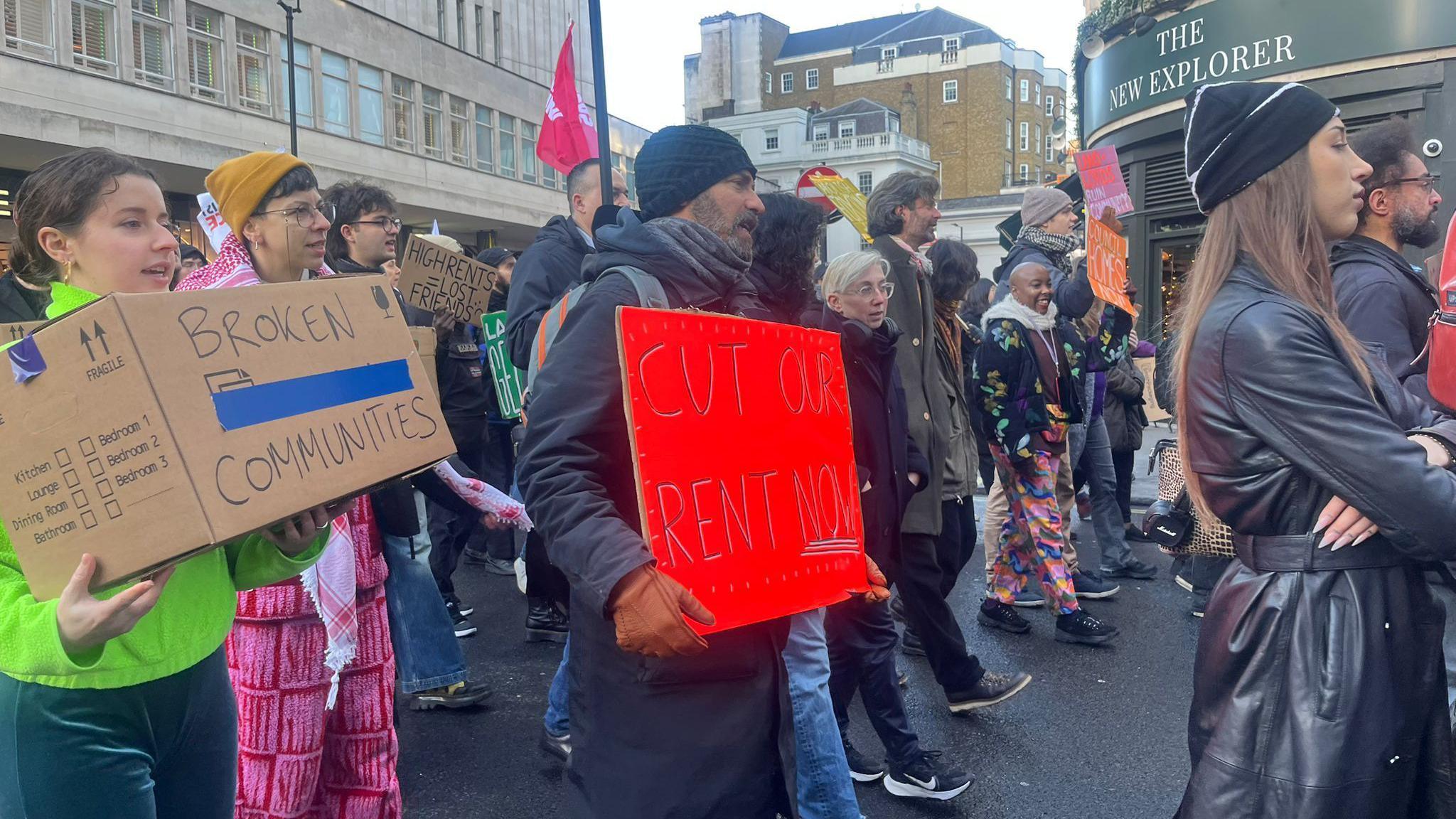 Protesters march with signs demanding rent cuts and highlighting "broken communities," and "cut our rent"