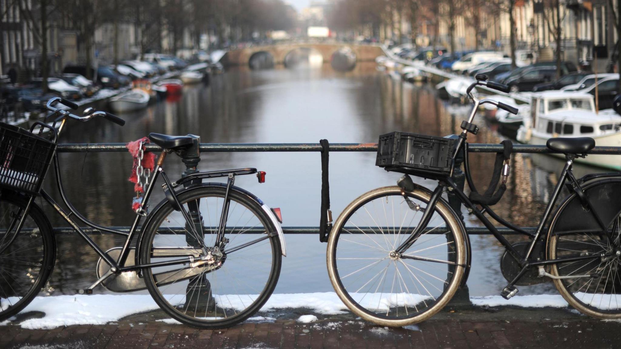 Two bicycles chained to an iron fence on a bridge. A canal lined with small boats is pictured beyond them.