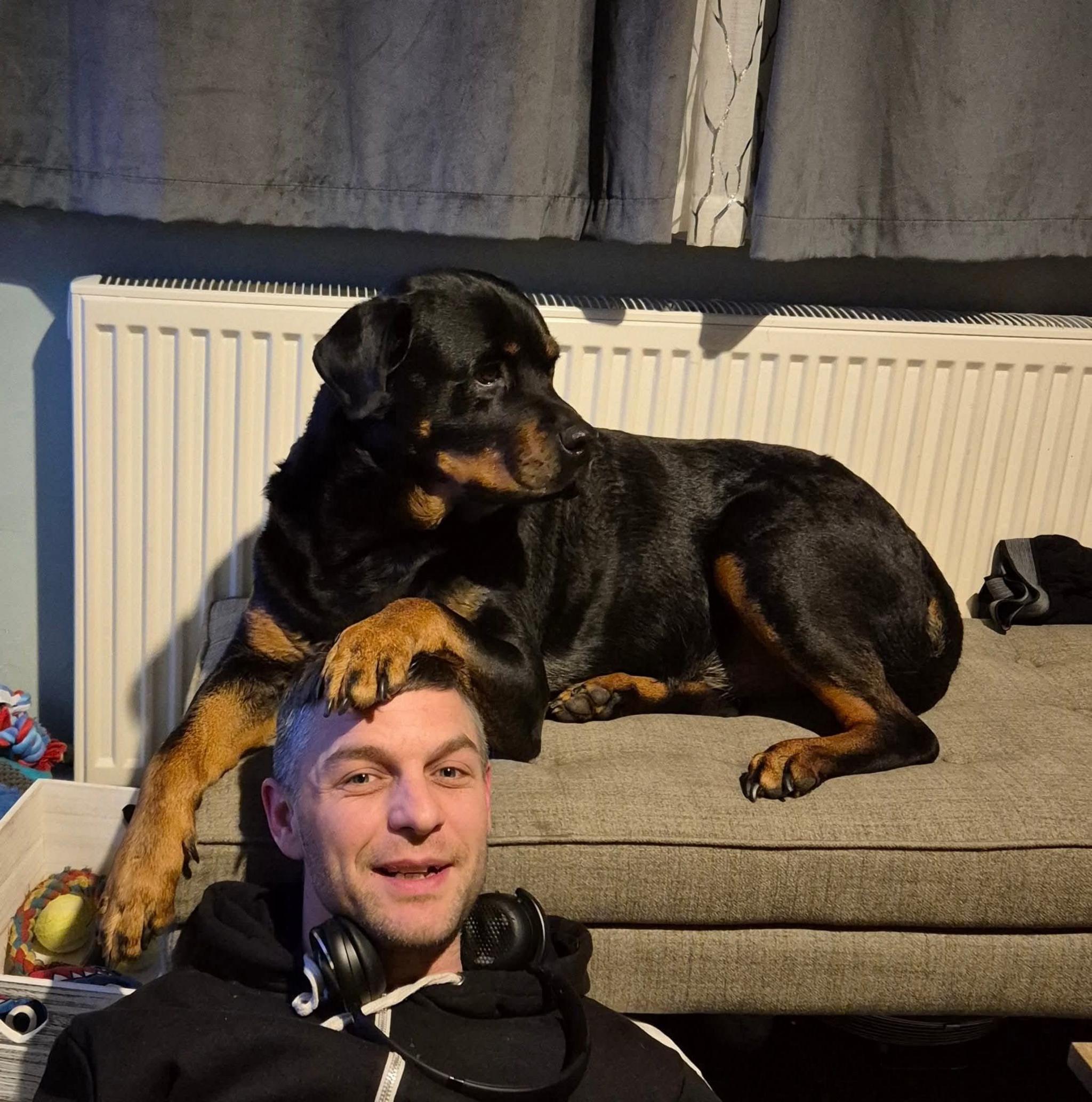 A man wearing a black jumper and headphones is sitting on the ground with the paw of a large black and brown dog, which is sitting on a raised padded seat, resting on his head. There is a radiator and curtains behind them.
