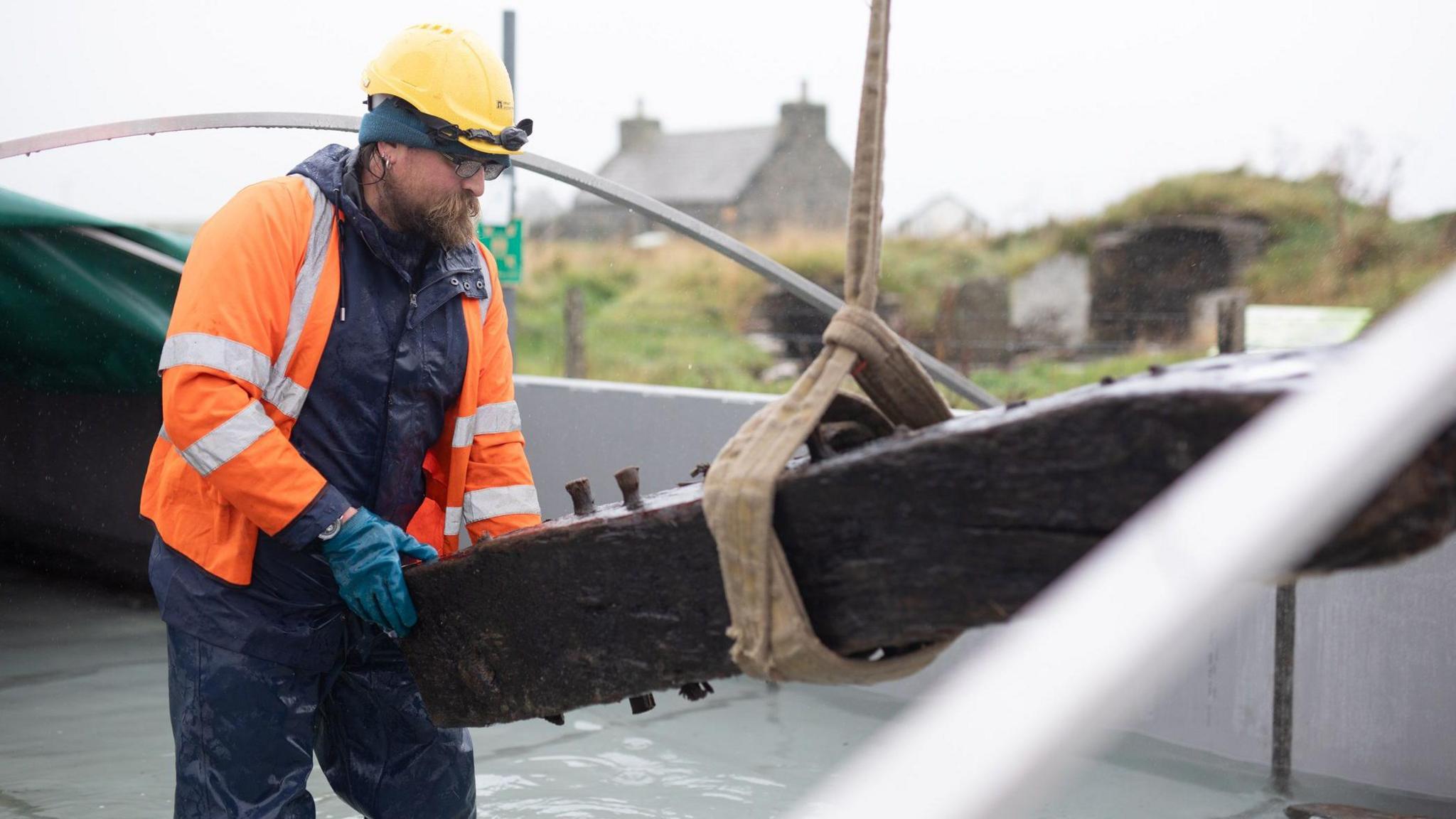 A bearded man in an orange hi vis jacket places a large timber beam into a water tank he is standing in