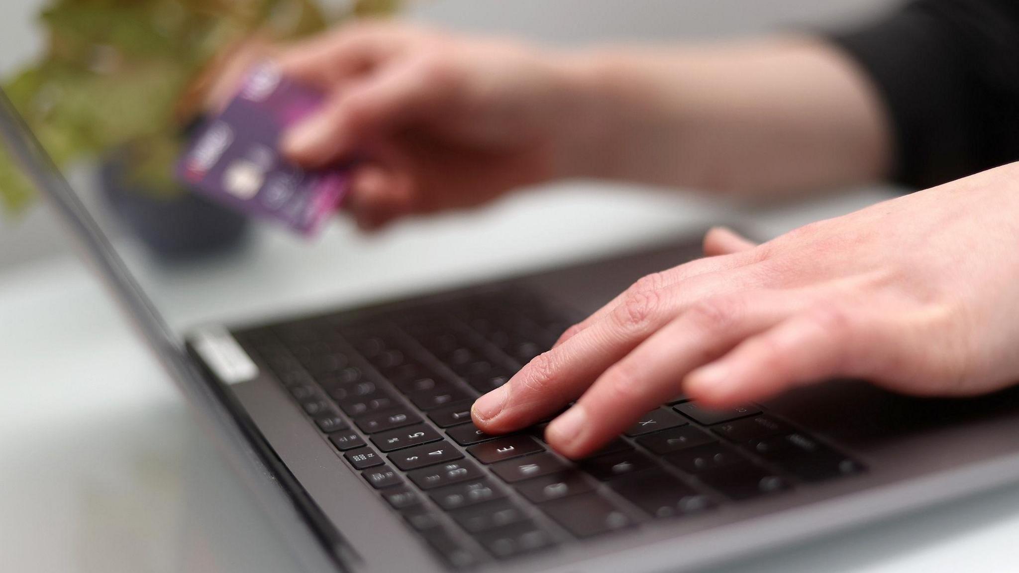 A close up of someone's hands with the left hand on the keys of a laptop and the right hand holding a bank card.