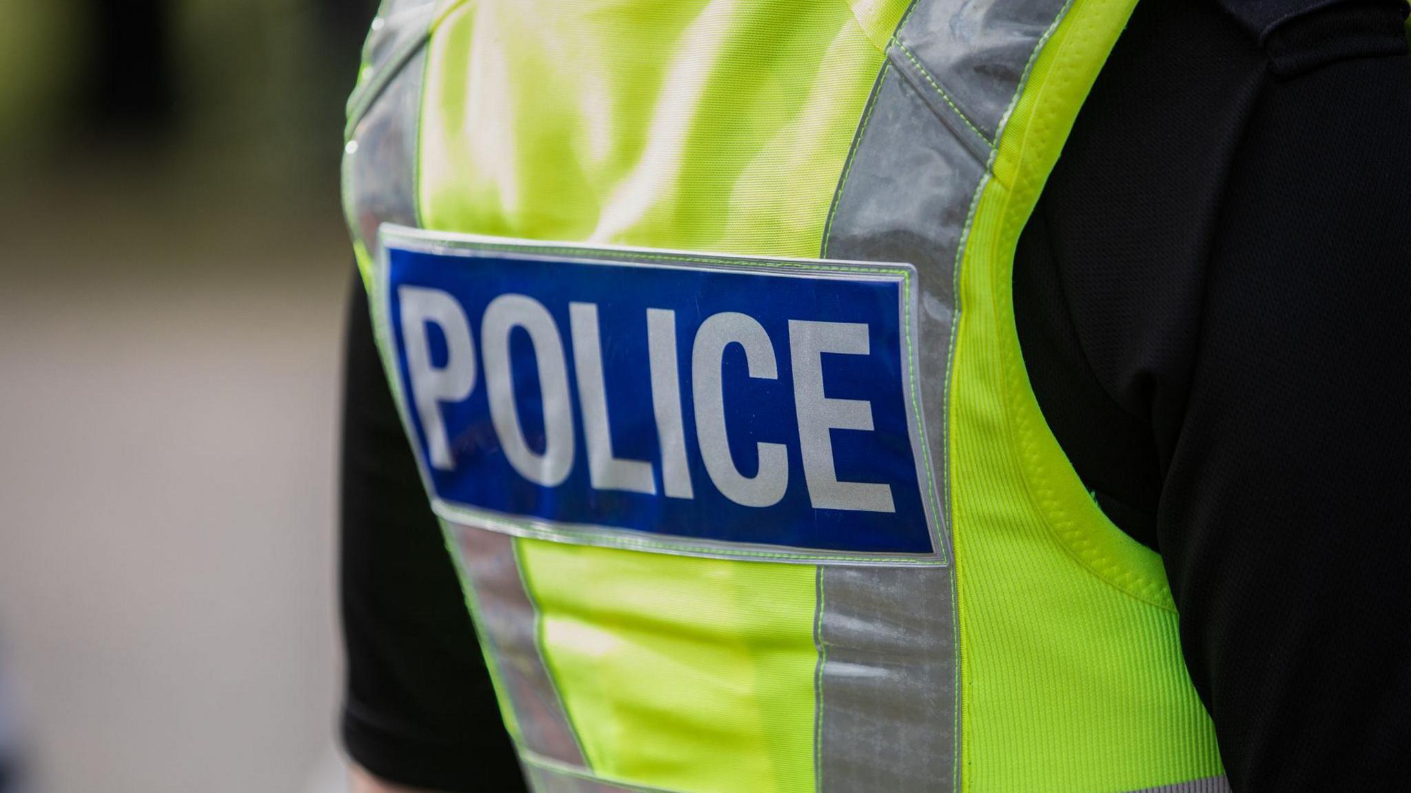 A photo of the back of a police officer in uniform. It shows a person wearing a black t-shirt with a yellow hi-vis police vest, with a blue and silver "POLICE" label on the back.