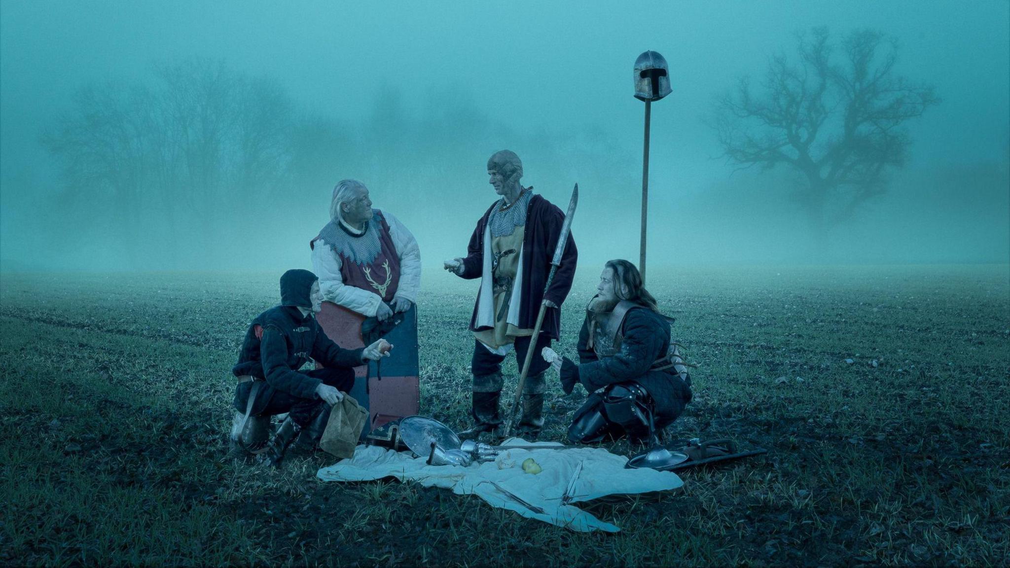 Four people dressed in medieval clothes are sitting on a field. It is dark and fog is coming up from the ground.