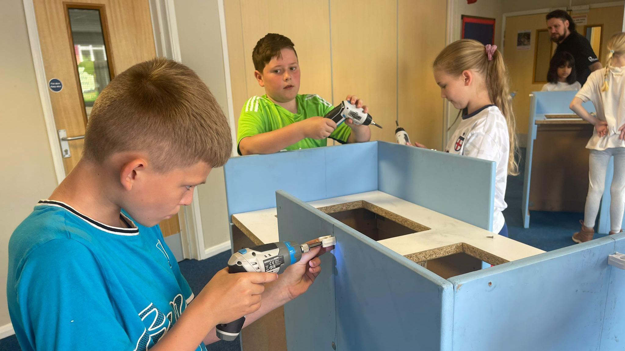 A child wearing a blue t-shirt drills a fitting into a wooden cabinet as another child holding a drill looks on.