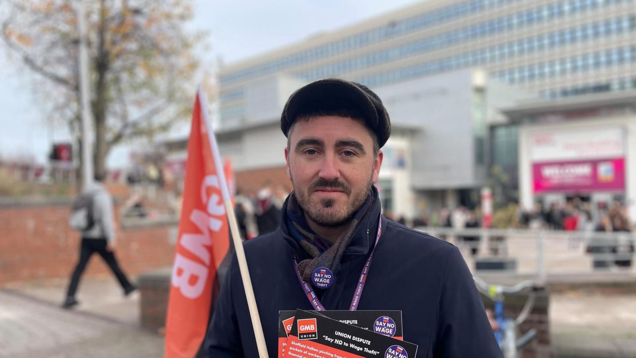 A man wearing a cap and gloves and holding a flag and several flyers, stands infront of a University building.