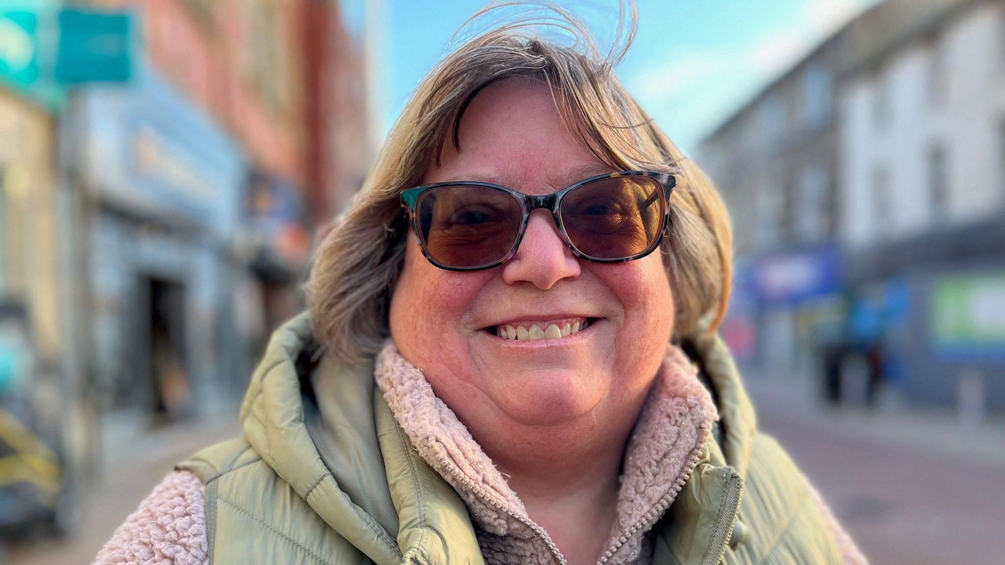 Valerie Long is pictured on the high street in Rhyl. She's wearing a light green gilet with a fluffy pink fleece. She is wearing sunglasses and has a light brown bob.